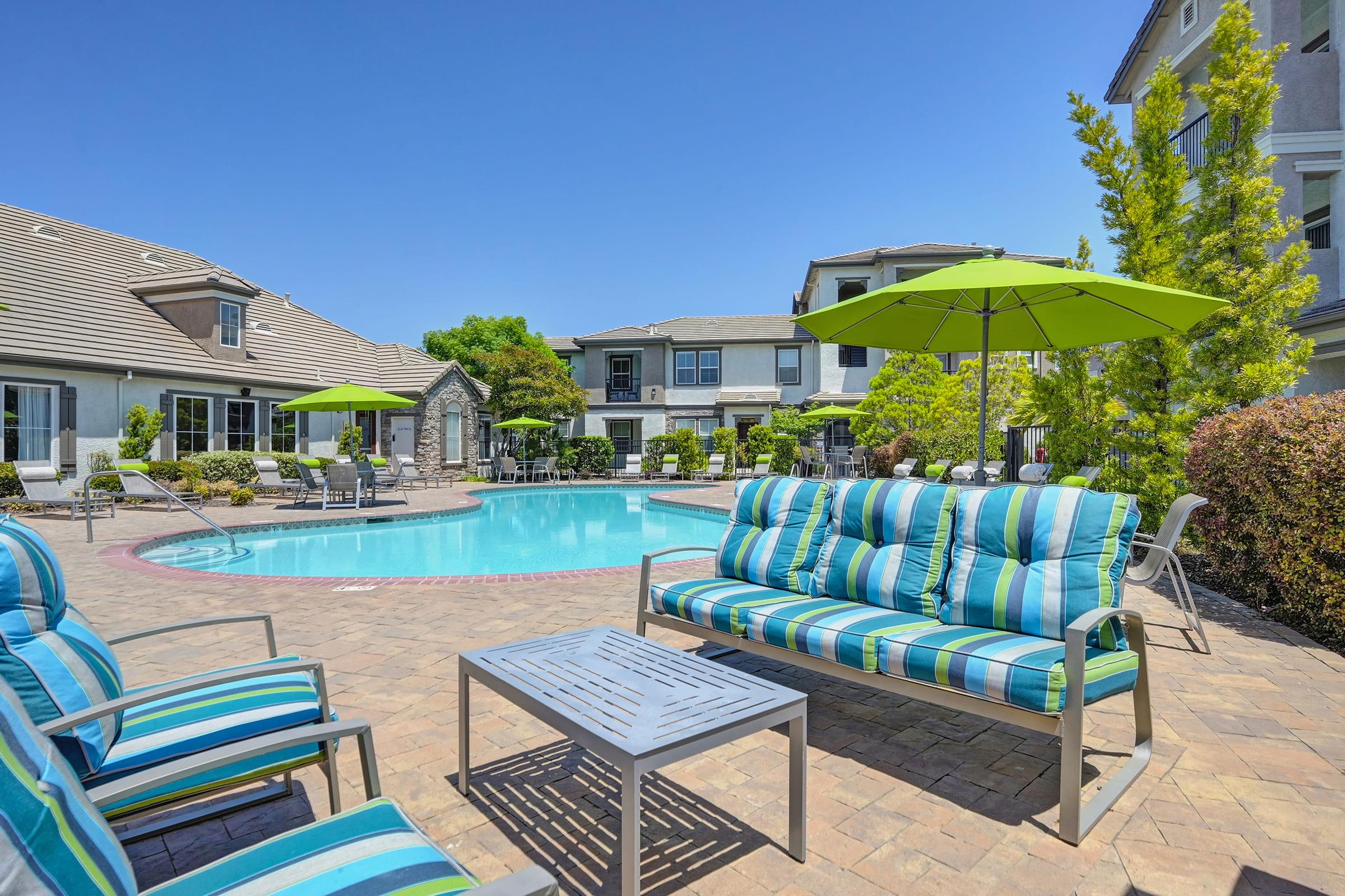 A picturesque pool area featuring a sparkling blue pool surrounded by comfortable lounge chairs and vibrant green umbrellas. In the foreground, striped outdoor seating is set around a small table, with lush greenery and modern buildings in the background under a clear blue sky.