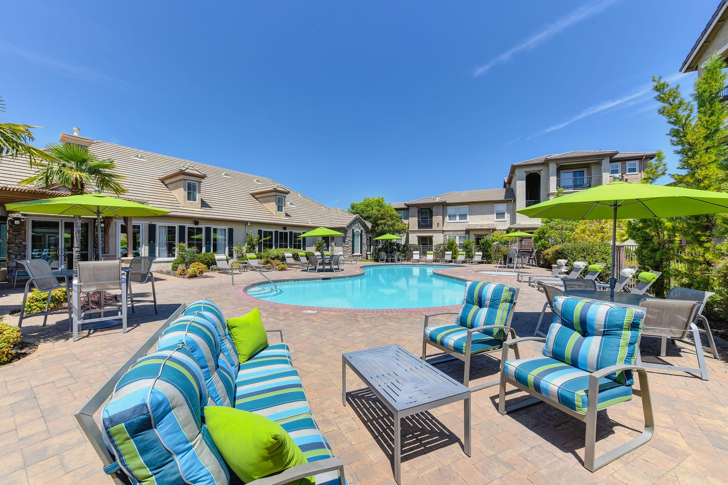 A sunny poolside area featuring a clear blue pool surrounded by lounge chairs and green umbrellas. Lush landscaping enhances the setting, with a stylish seating arrangement in blue and green stripes. The backdrop includes modern buildings, creating a relaxed, inviting atmosphere.