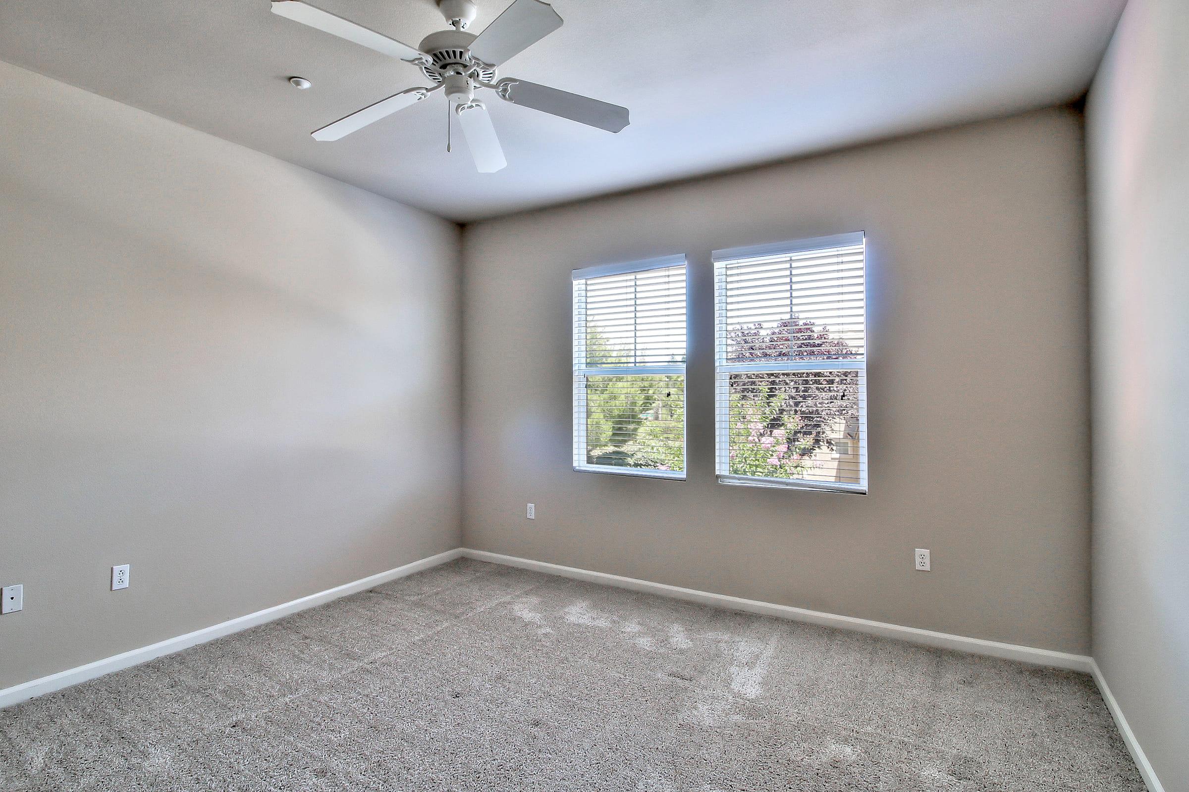 A vacant room featuring light beige walls and a ceiling fan. Two windows with white blinds allow natural light to enter, showcasing greenery outside. The floor is covered with light gray carpet, and there are no furnishings or decorations present, giving the space a clean and open appearance.