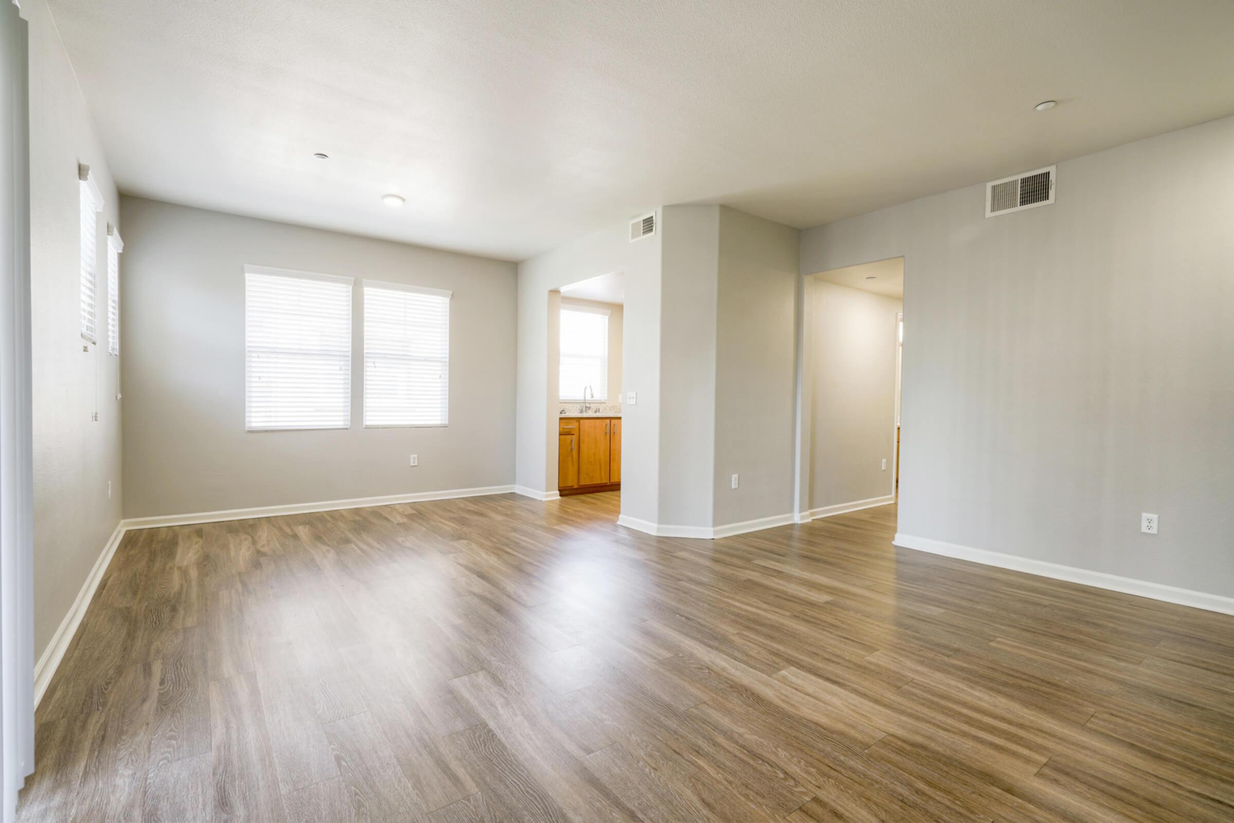 Spacious room with light-colored walls and wood-like flooring. Large windows on one wall allow natural light to fill the space. A doorway leads to a kitchen area with wooden cabinets visible, while another doorway opens to a hallway. The overall atmosphere is bright and inviting.