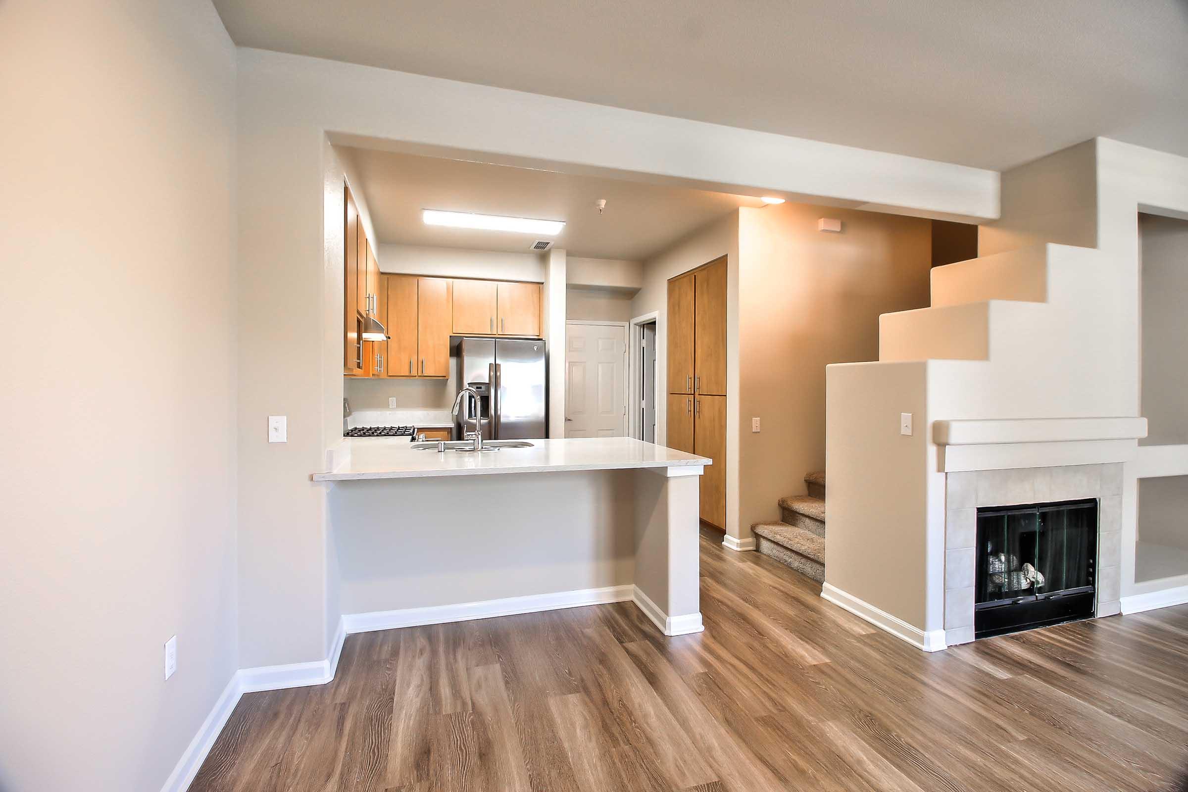 Modern kitchen and living area featuring wooden cabinetry, stainless steel appliances, and a countertop bar. A staircase leads to a second level, and there's a small fireplace integrated into the wall. The flooring is hardwood, creating a warm and inviting atmosphere.