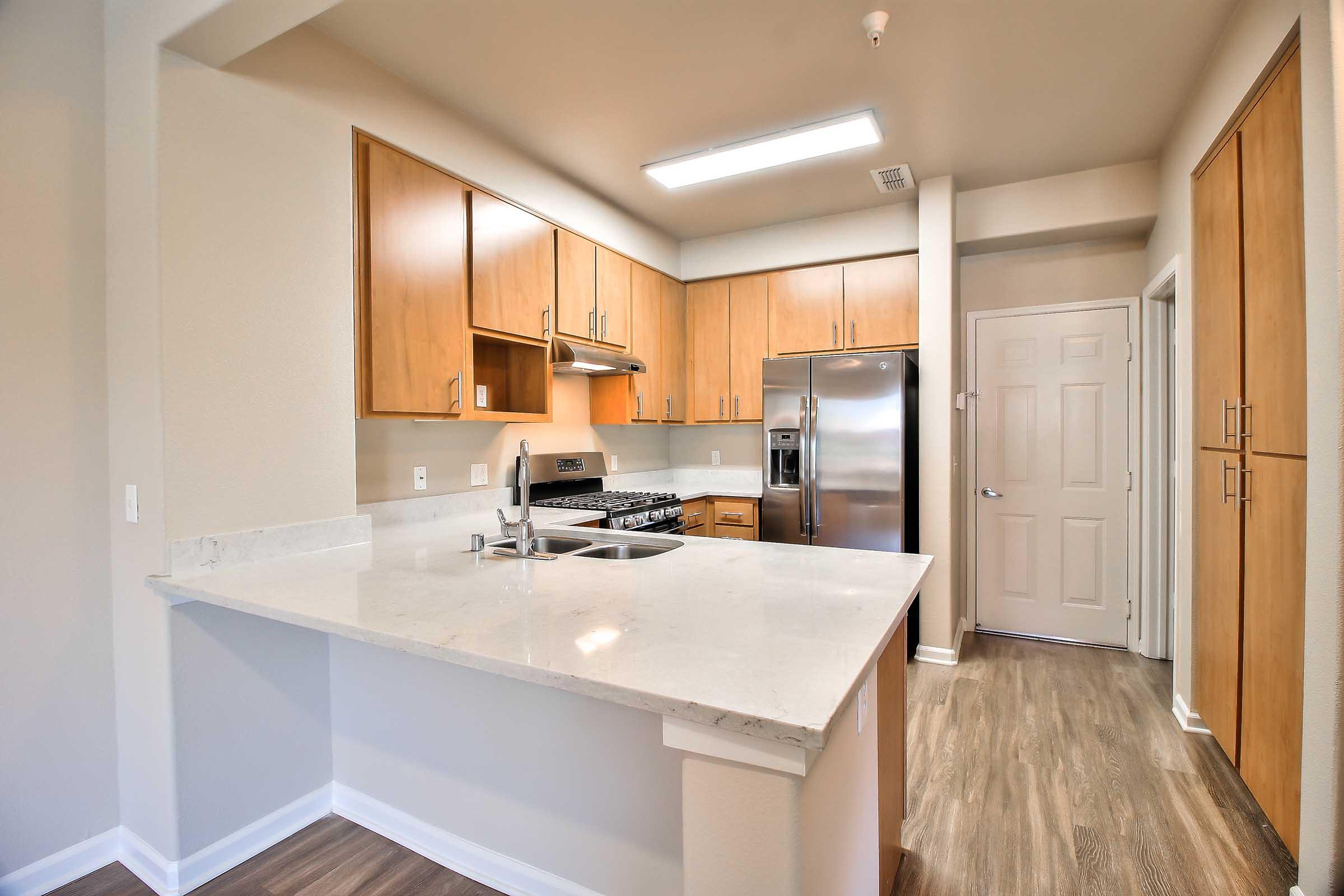 Modern kitchen featuring wooden cabinetry, stainless steel appliances, a large countertop with a sink, and an open layout. The space is illuminated by overhead lighting, with a door leading to another room in the background. The flooring is a light wood look, creating a warm and inviting atmosphere.