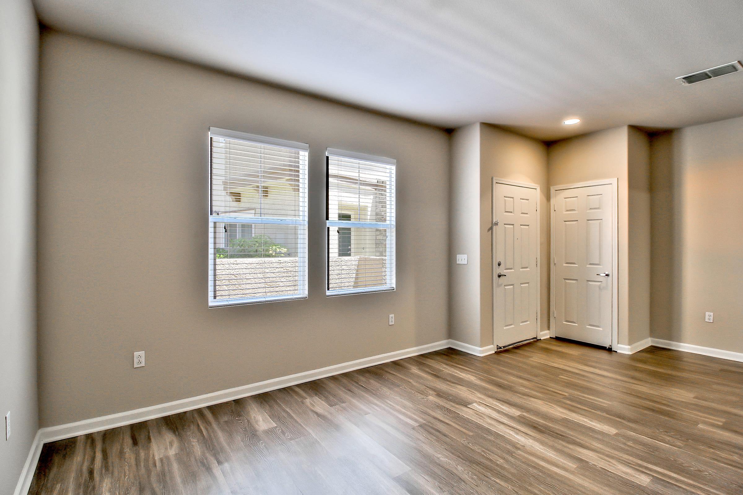 A spacious, empty room with light-colored walls and wood-like flooring. Two windows allow natural light to enter, and there are two doors leading to exits. The overall atmosphere is bright and inviting, suitable for various interior designs.