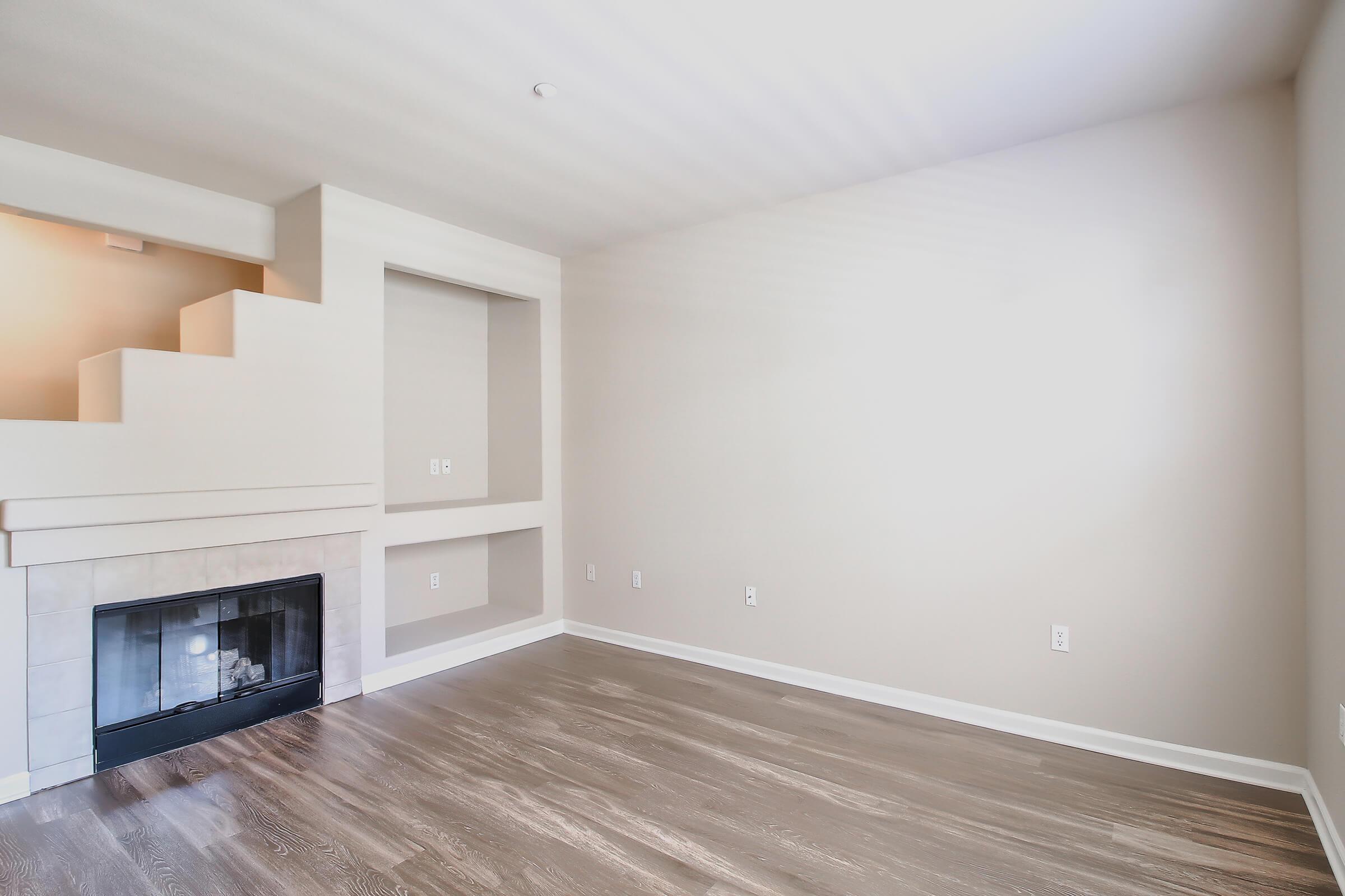 A spacious, empty living room featuring a modern fireplace, light-colored walls, and hardwood flooring. There is a staircase on the left side leading to an upper level, and a small recessed area built into the wall, providing a minimalist and inviting atmosphere.