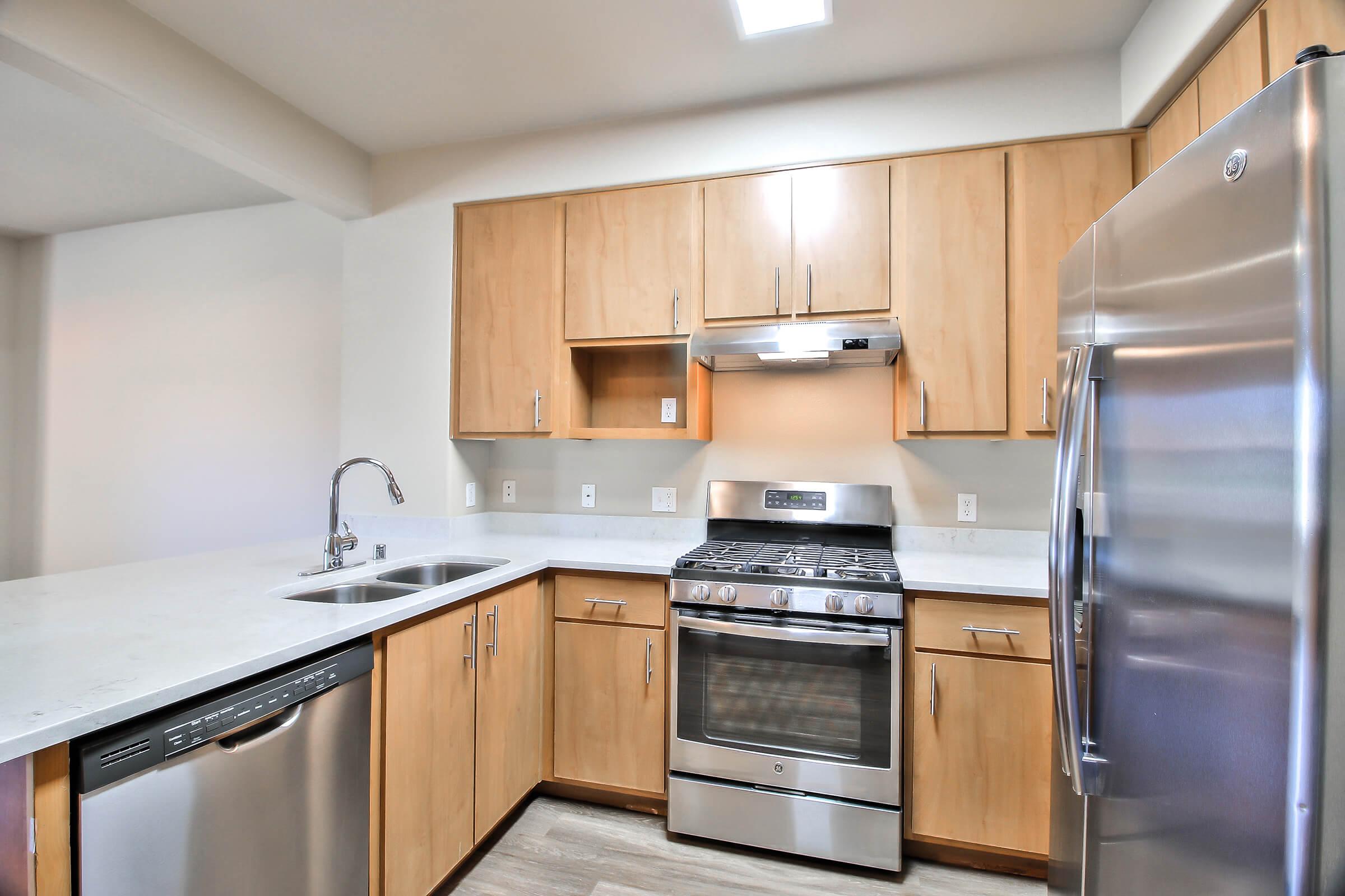 Modern kitchen featuring light wood cabinetry, a stainless steel refrigerator, a gas range with an overhead exhaust, and a double-basin sink. The countertops are light-colored, and the space is well-lit with natural light and overhead fixtures.