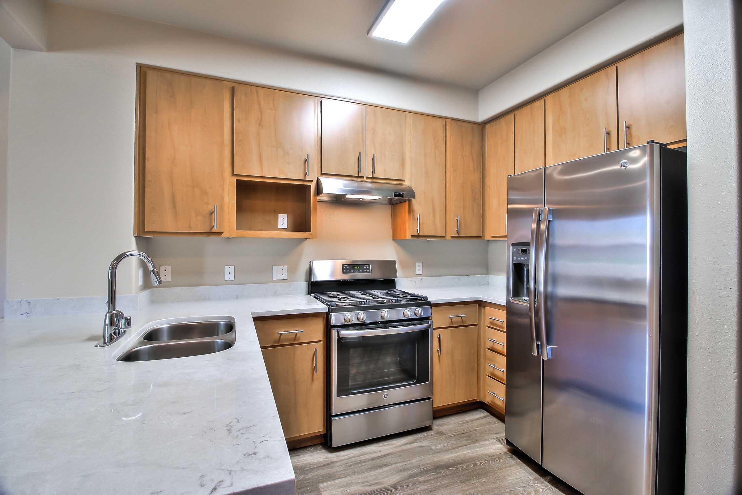 Modern kitchen featuring light wood cabinets, stainless steel appliances including a gas stove and refrigerator, a double sink, and marble-style countertops. The space is well-lit with overhead lighting and has a clean, contemporary design.
