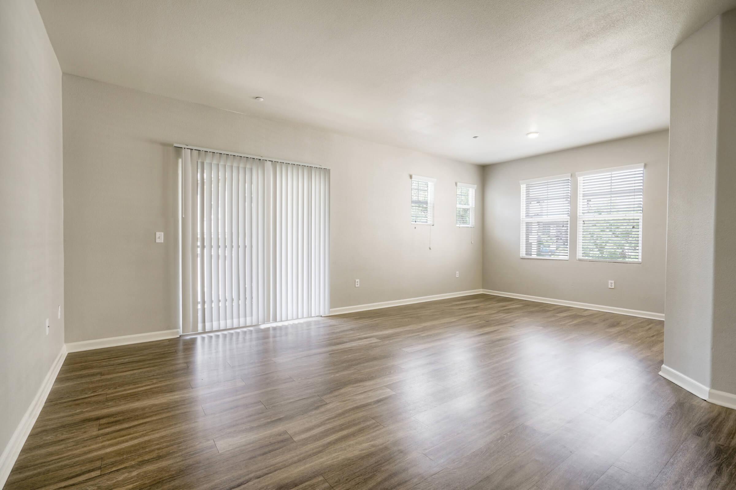 A spacious, empty living room with light-colored walls and brown wooden flooring. To the left, there are sliding glass doors with vertical blinds that open to an outdoor area. Natural light enters through multiple windows, creating a bright and airy atmosphere.