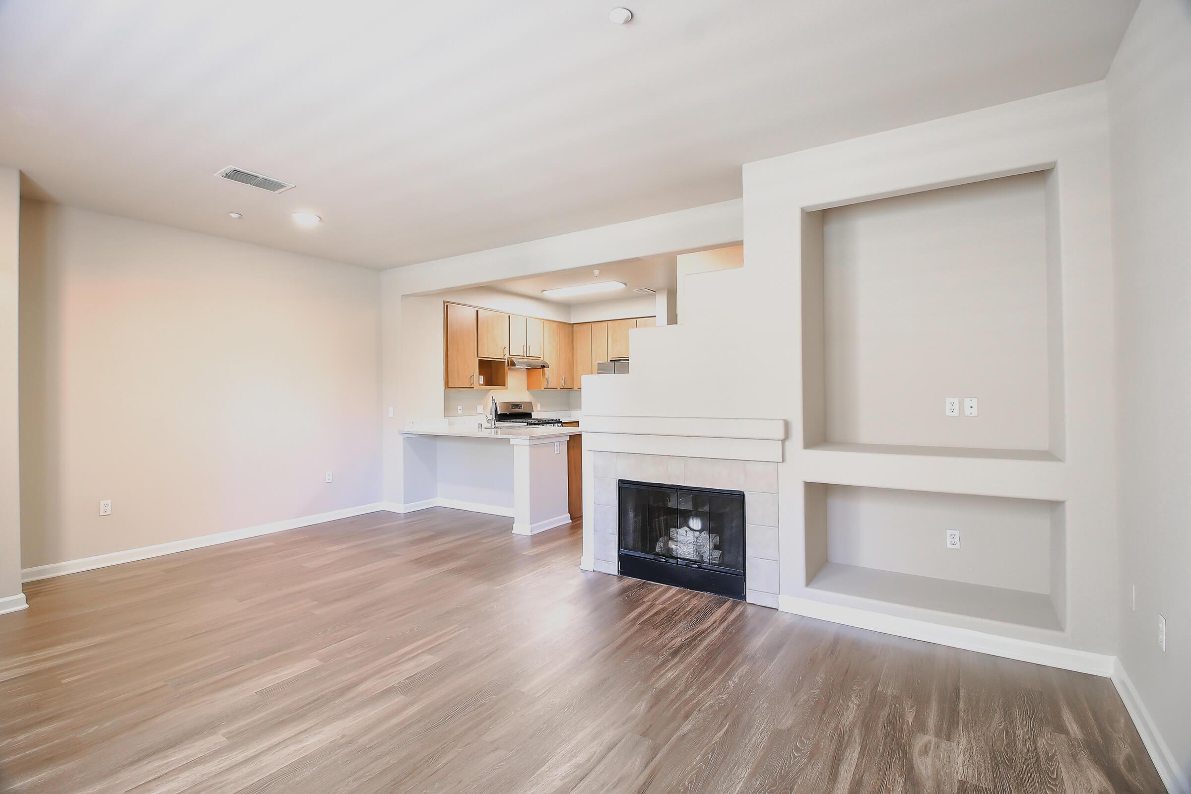 Spacious living room with hardwood floors, featuring a white fireplace and built-in shelving. The area connects to a modern kitchen with wooden cabinets, creating an open-concept layout. Bright natural light fills the space from multiple windows.