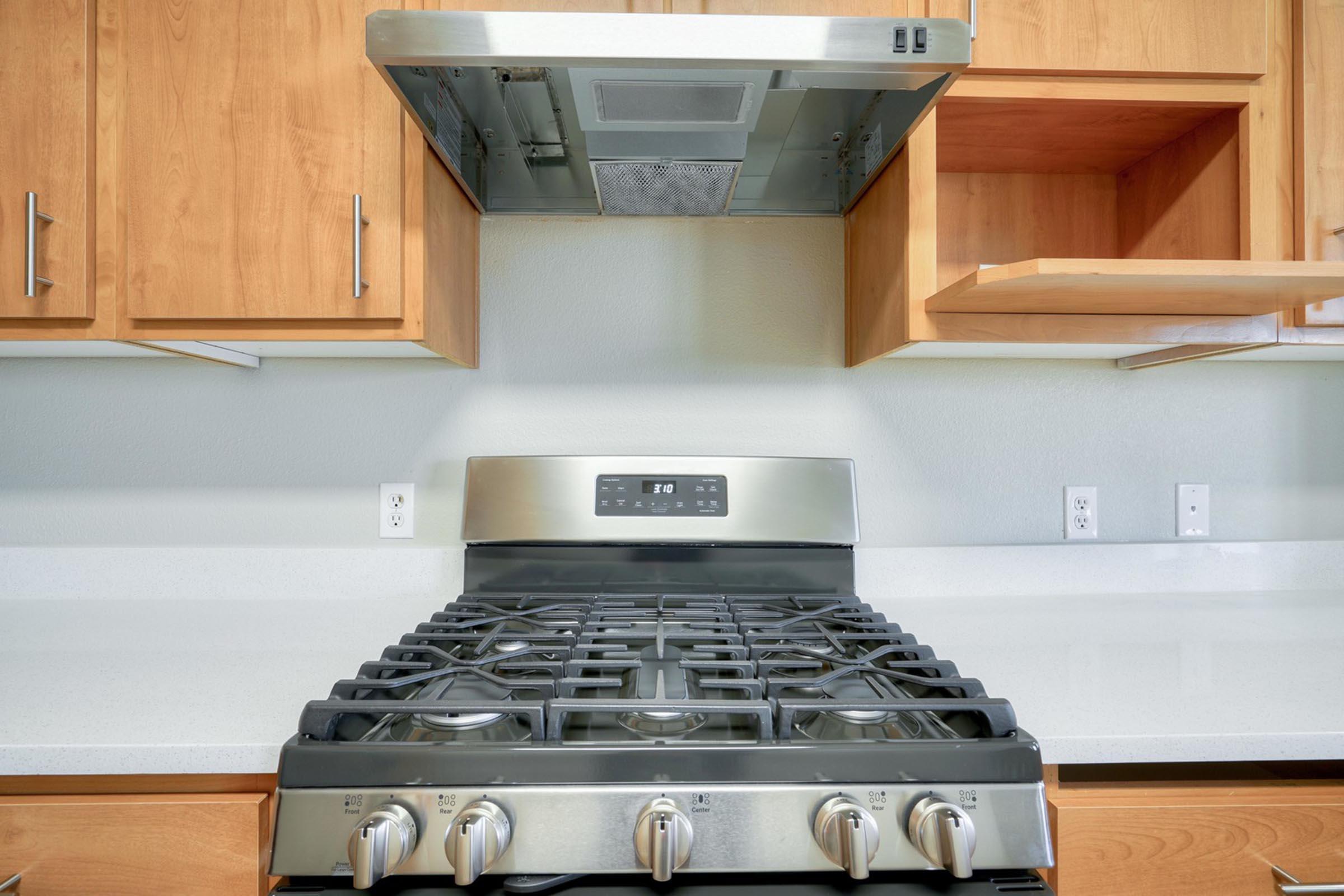 Modern kitchen featuring a stainless steel gas stove with multiple burners, below a sleek range hood mounted above. Wooden cabinetry in a light finish complements the clean, contemporary design of the countertop, creating a functional and stylish cooking space.