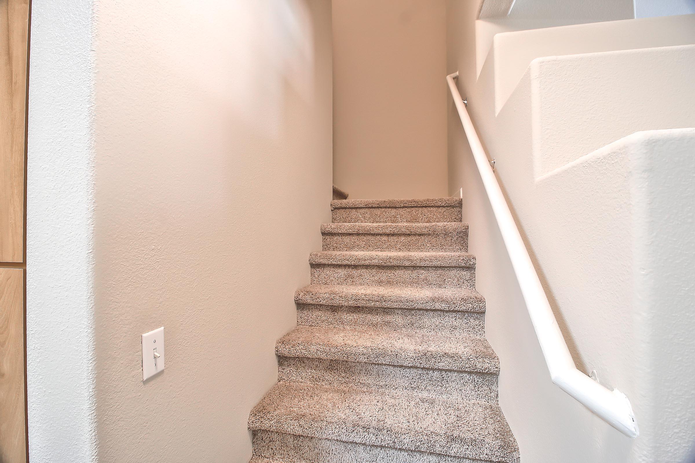 A well-lit staircase with beige carpeting, leading upward. The walls are painted a soft neutral color, and a white handrail runs alongside the steps. The design is simple and modern, creating a clean and inviting atmosphere.