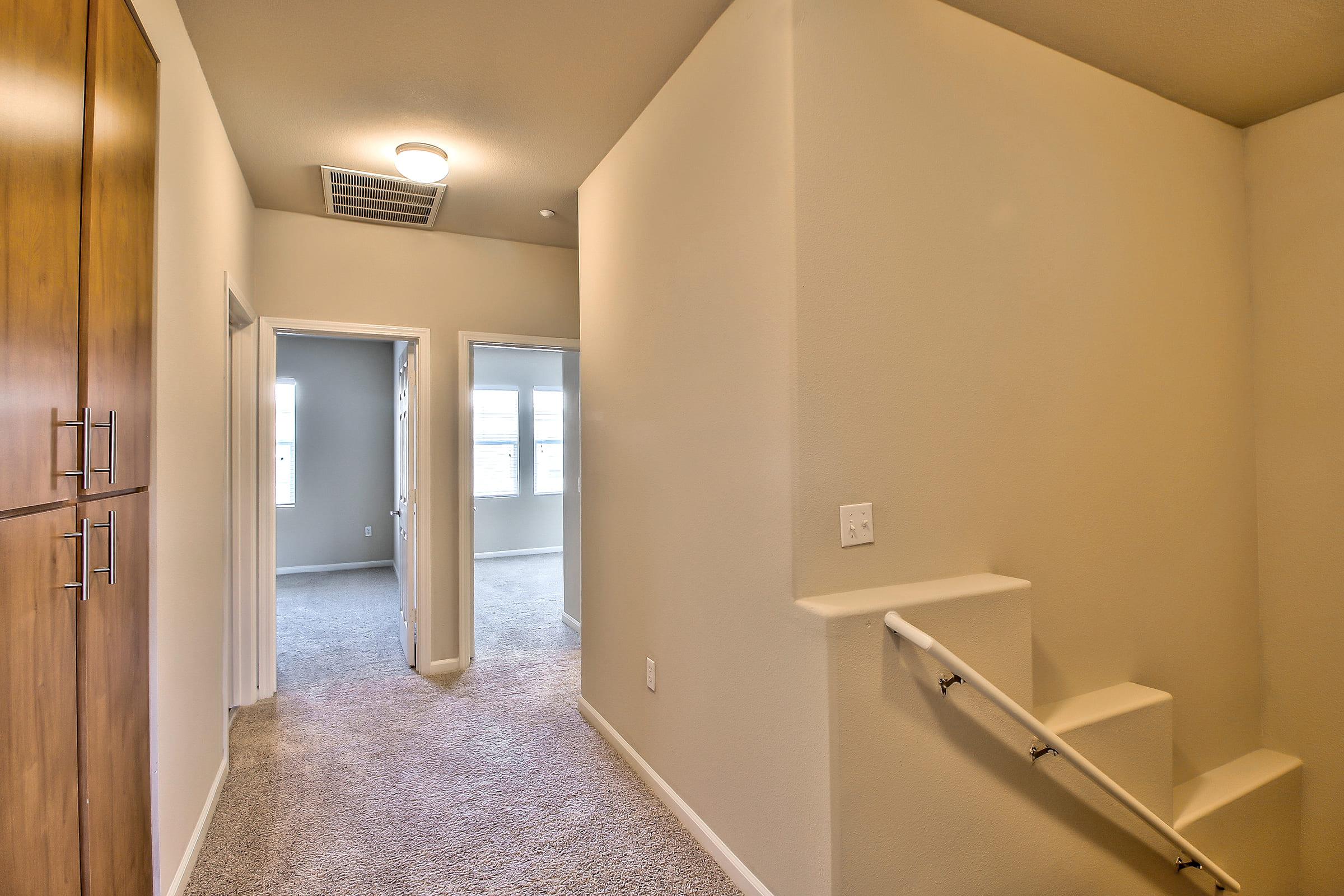 A well-lit hallway featuring beige walls and carpeted flooring. On the left, there is a wooden cabinet. To the right, a staircase leads to an upper level. Two open doorways at the end of the hallway lead to additional rooms with windows providing natural light.