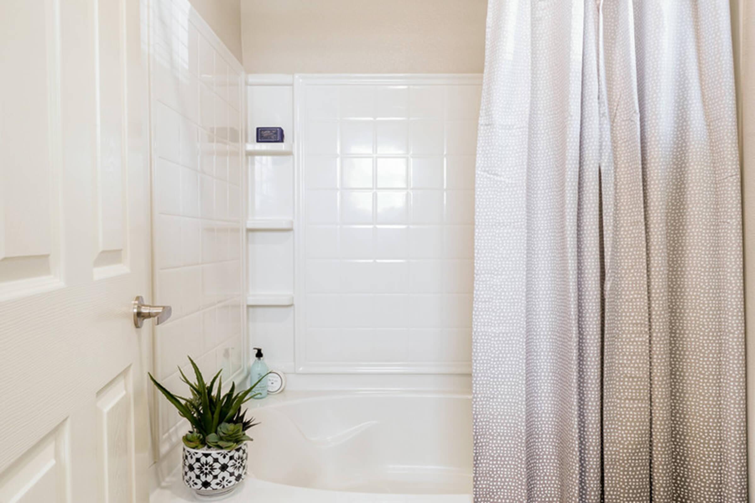 A clean and bright bathroom featuring a bathtub with a shower curtain, a built-in shelf with toiletries, and a decorative plant in a patterned pot. The walls are tiled with white tiles, and natural light illuminates the space.