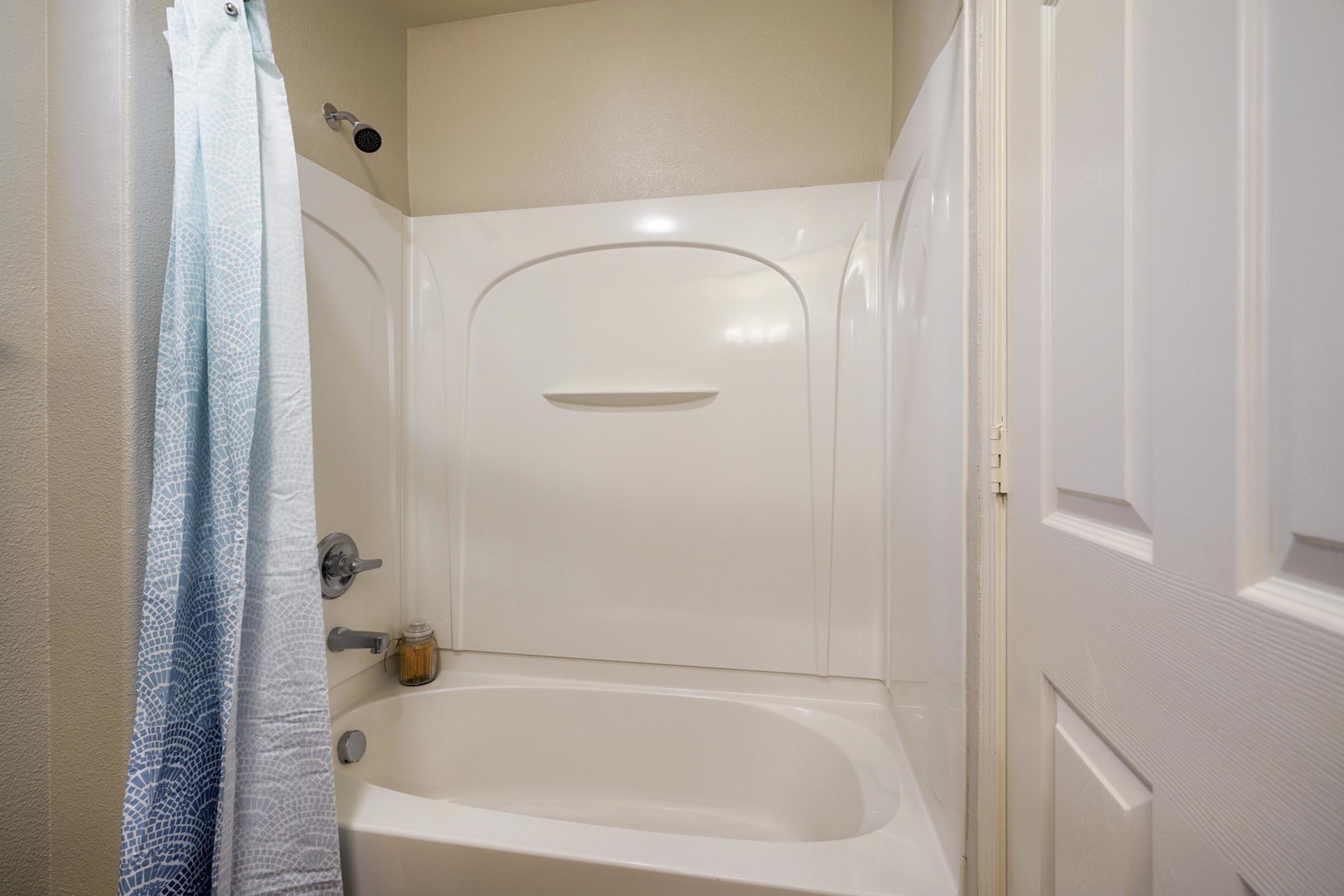 A clean bathroom with a white bathtub and a shower curtain draped to one side. The walls are a light color, and there is a small shelf or ledge in the tub area. A door is partially visible on the right, adding to the simple yet functional design of the space.