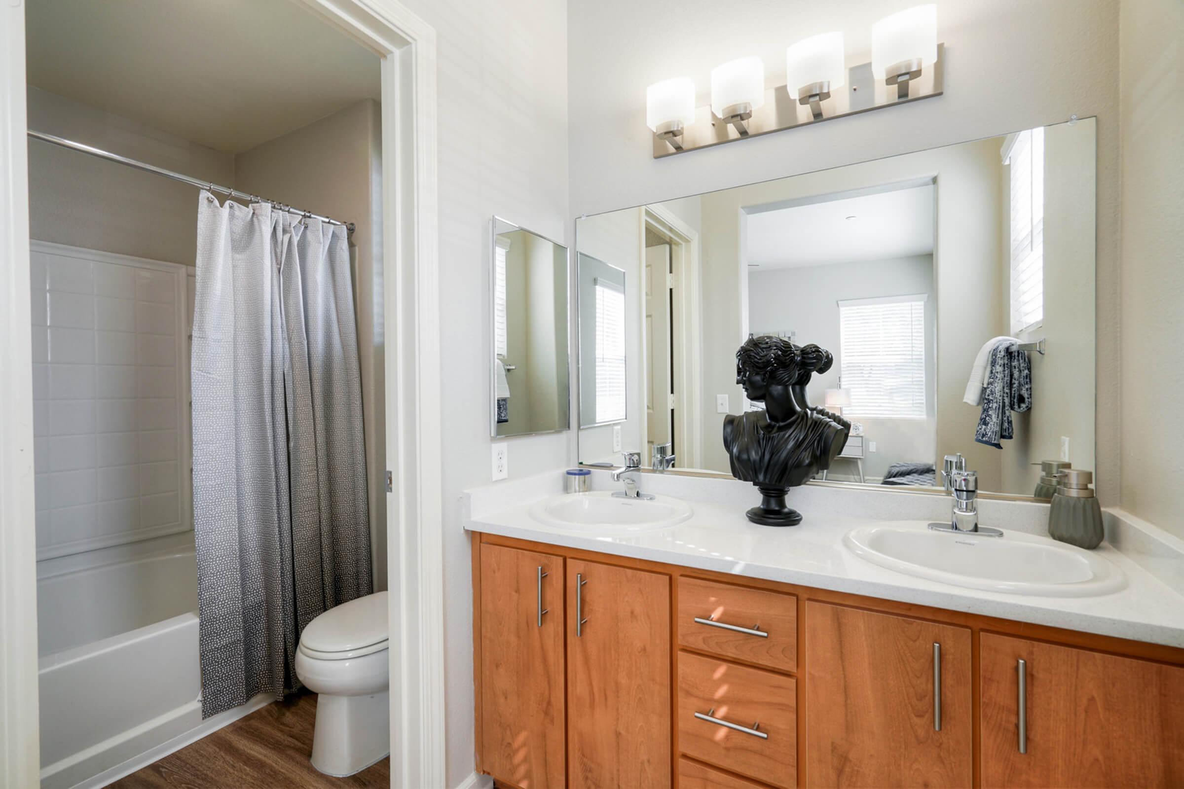 Modern bathroom featuring a double vanity with a large mirror, two sinks, and wooden cabinetry. A shower area with a curtain is visible, along with a toilet. Natural light streams in through a window, and a decorative bust adds a stylish touch to the décor.