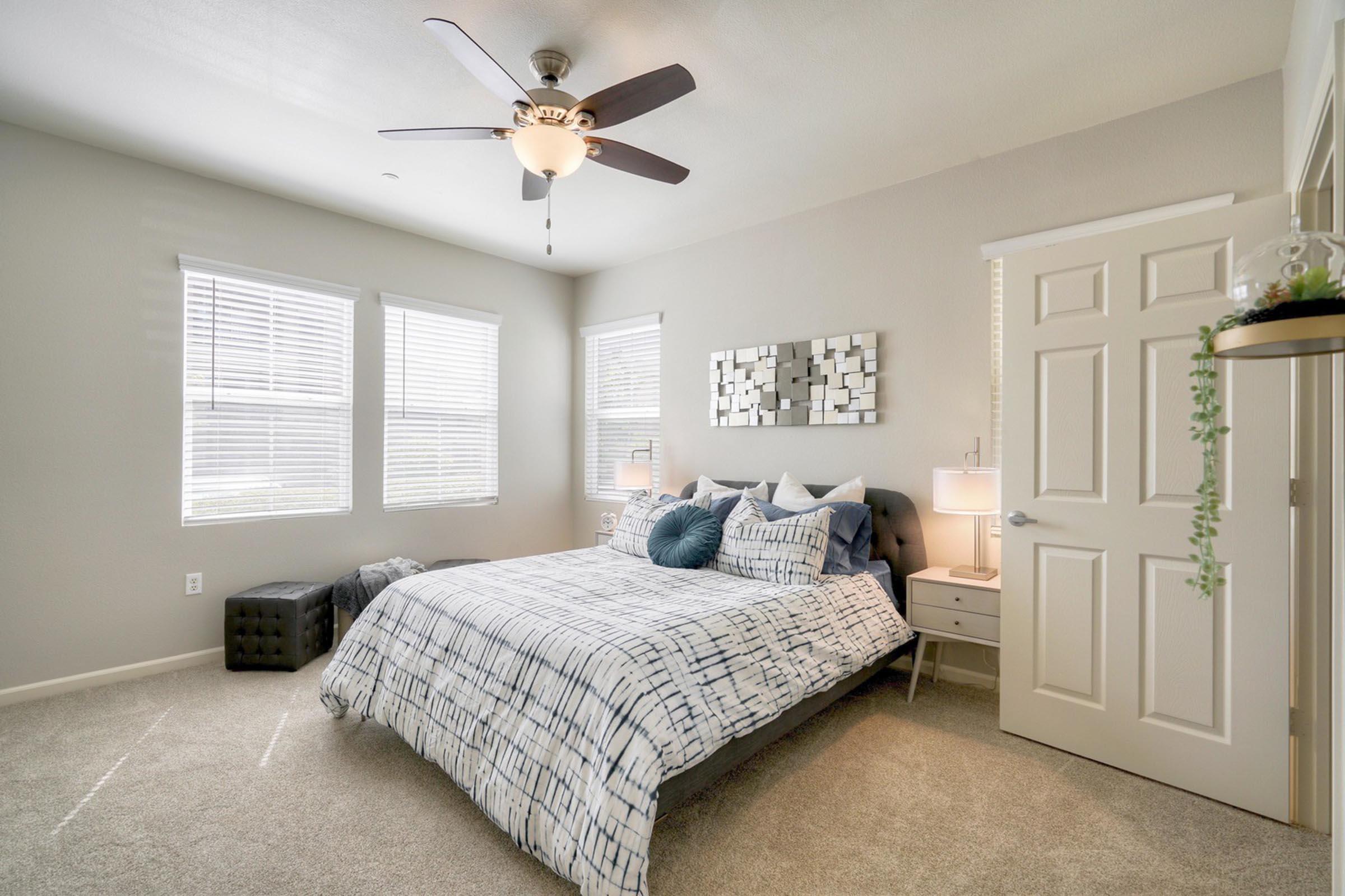 A cozy bedroom featuring a queen-sized bed with patterned bedding, two bedside tables with lamps, and a modern ceiling fan. Natural light streams in through multiple windows with blinds, and there's a decorative mirror on the wall. Soft carpet adds warmth to the space, enhancing its tranquil ambiance.