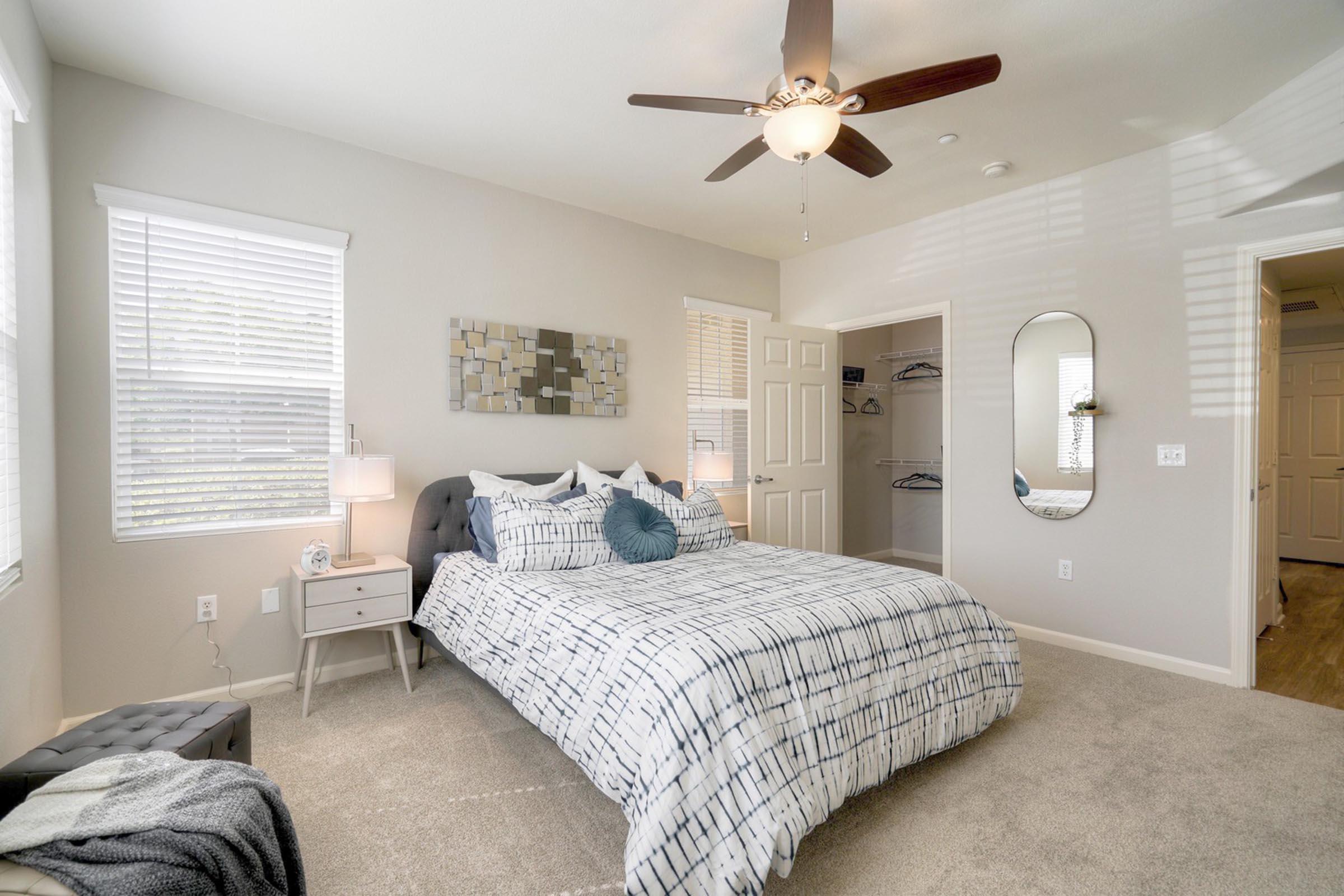 A cozy bedroom featuring a king-sized bed with patterned bedding, two bedside tables with lamps, a ceiling fan, and a large mirror. The walls are painted in light colors with ample natural light coming through the windows. A closet is visible in the background, and the carpet is soft and plush.