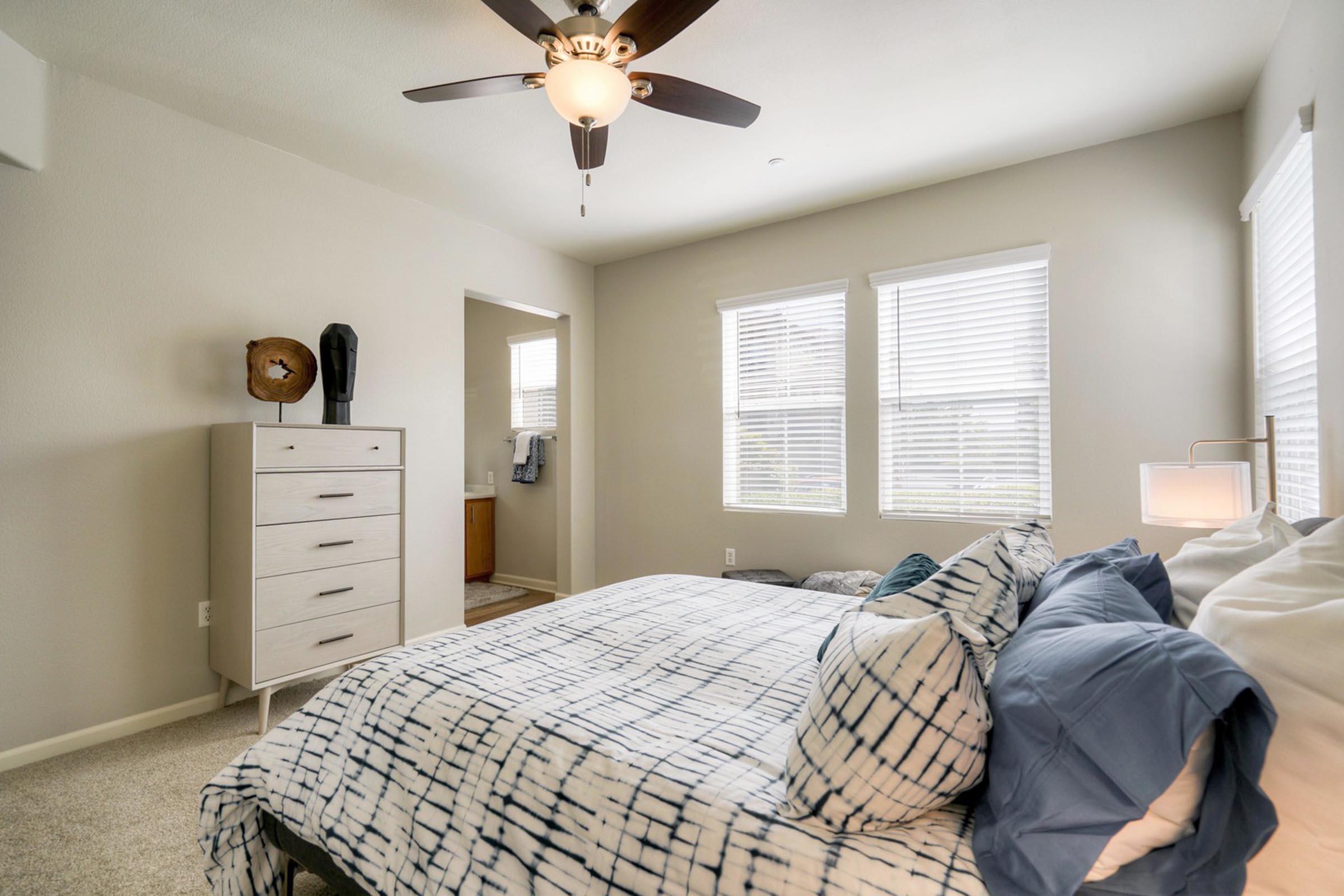 A cozy bedroom featuring a large bed with decorative pillows, a ceiling fan, and natural light coming through two windows. A white dresser is positioned against one wall, with a round clock and a black decorative object on top. A doorway leads to a bathroom area in the background.