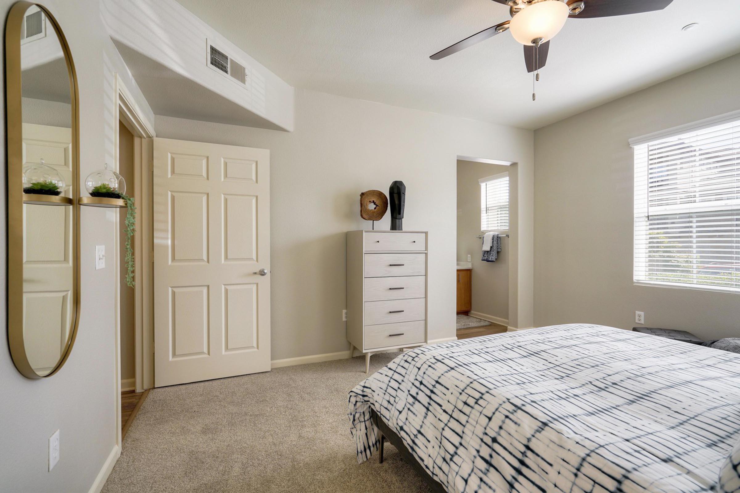 A cozy bedroom featuring light gray walls, a bed with a patterned duvet, a large mirror on the wall, a white dresser, and a doorway leading to an adjacent bathroom. Natural light streams through a window, enhancing the inviting atmosphere.
