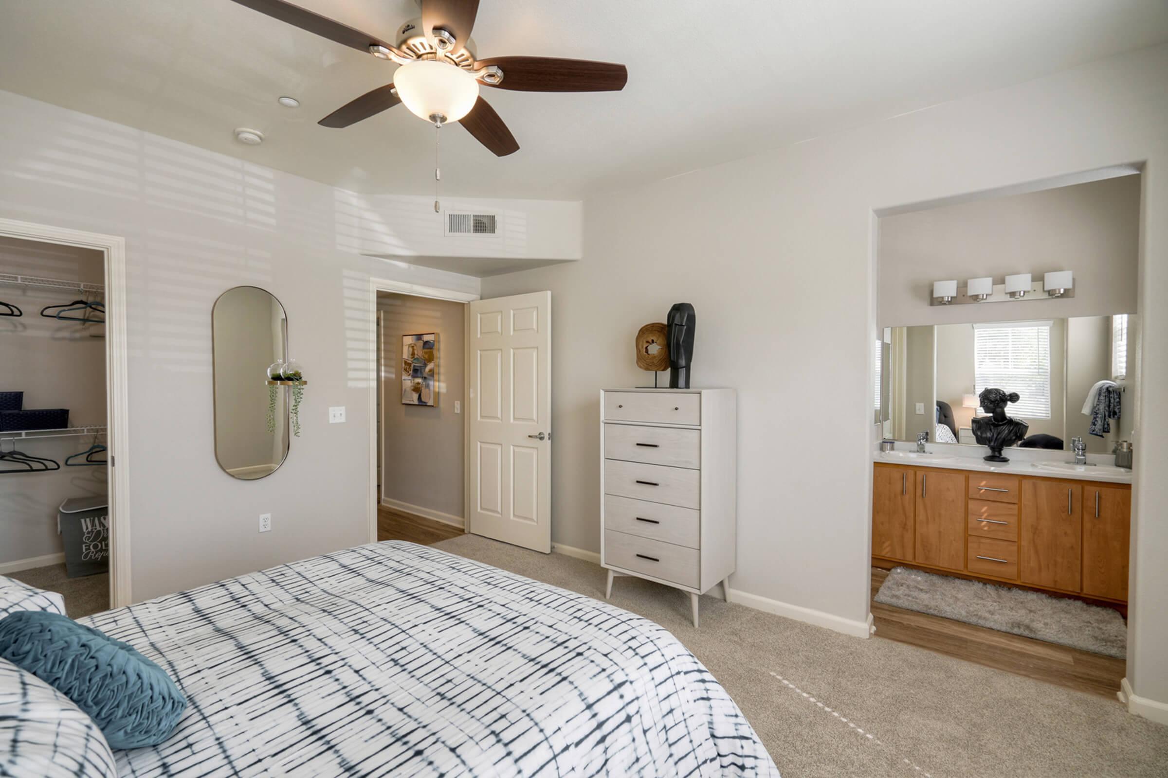 A bright bedroom with a bed featuring a patterned comforter. There's a ceiling fan, a mirror on the wall, and a closet with hanging space visible. A bathroom is through a doorway, showing a double vanity and large mirror. The carpeted floor adds a cozy touch to the space.