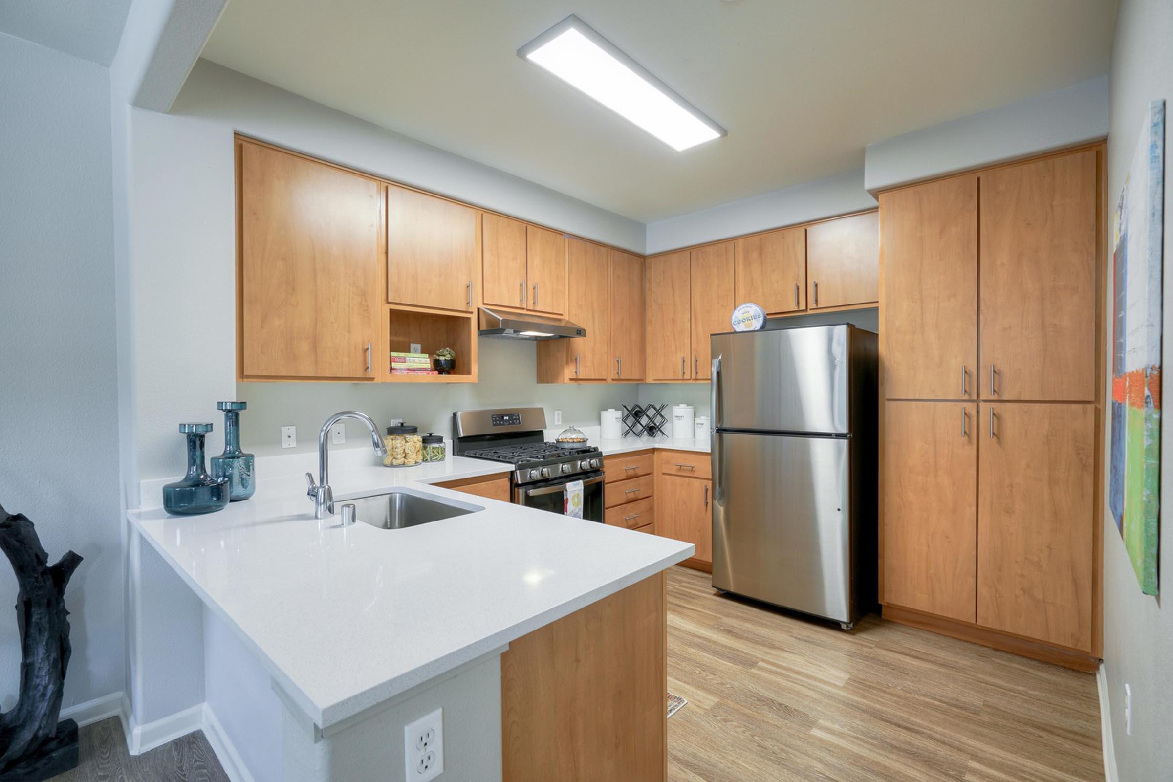 Modern kitchen with wooden cabinetry, stainless steel appliances including a refrigerator and stove, and white quartz countertops. The space is well-lit with overhead lighting and features decorative items on the shelves, alongside a sink and a small island bar. The floor is covered in light wooden laminate.