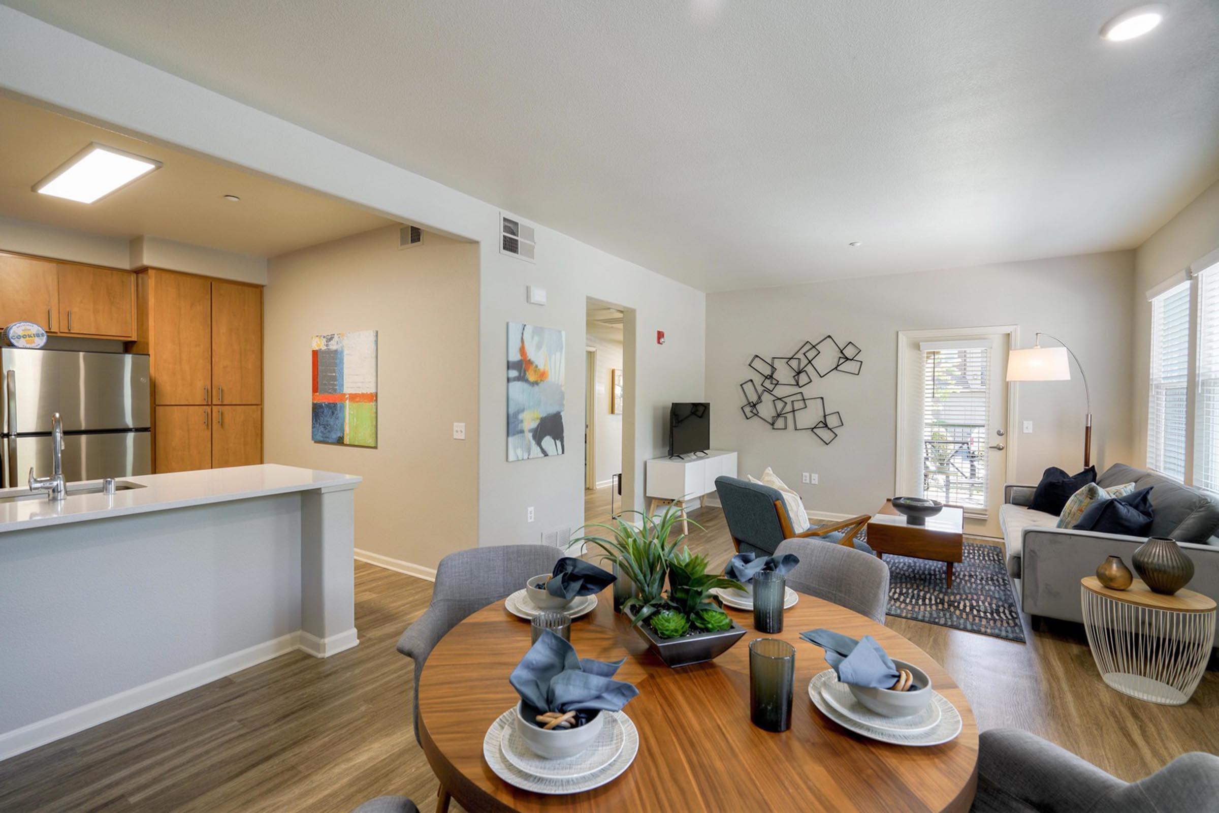 Bright and modern open-concept living space featuring a dining area with a round wooden table set for four, contemporary kitchen with stainless steel appliances, cozy seating area with a gray couch and decorative pillows, and stylish decor throughout. Large windows allow natural light to fill the room.