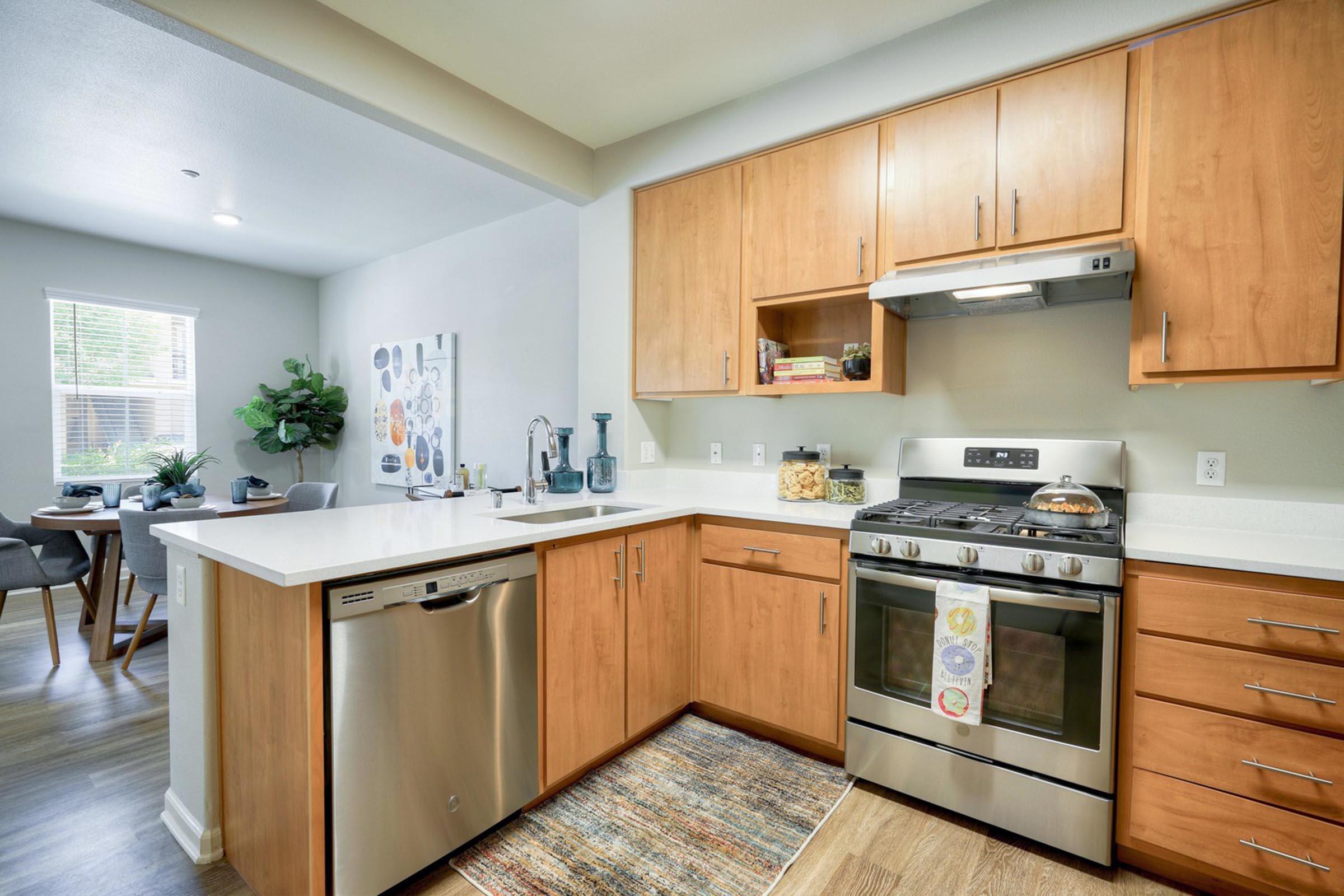 Modern kitchen featuring wooden cabinets, stainless steel appliances including a gas stove and dishwasher, and a white countertop. A colorful kitchen towel hangs from the stove. In the background, a dining area with a table and chairs is visible, alongside a wall adorned with decorative art.