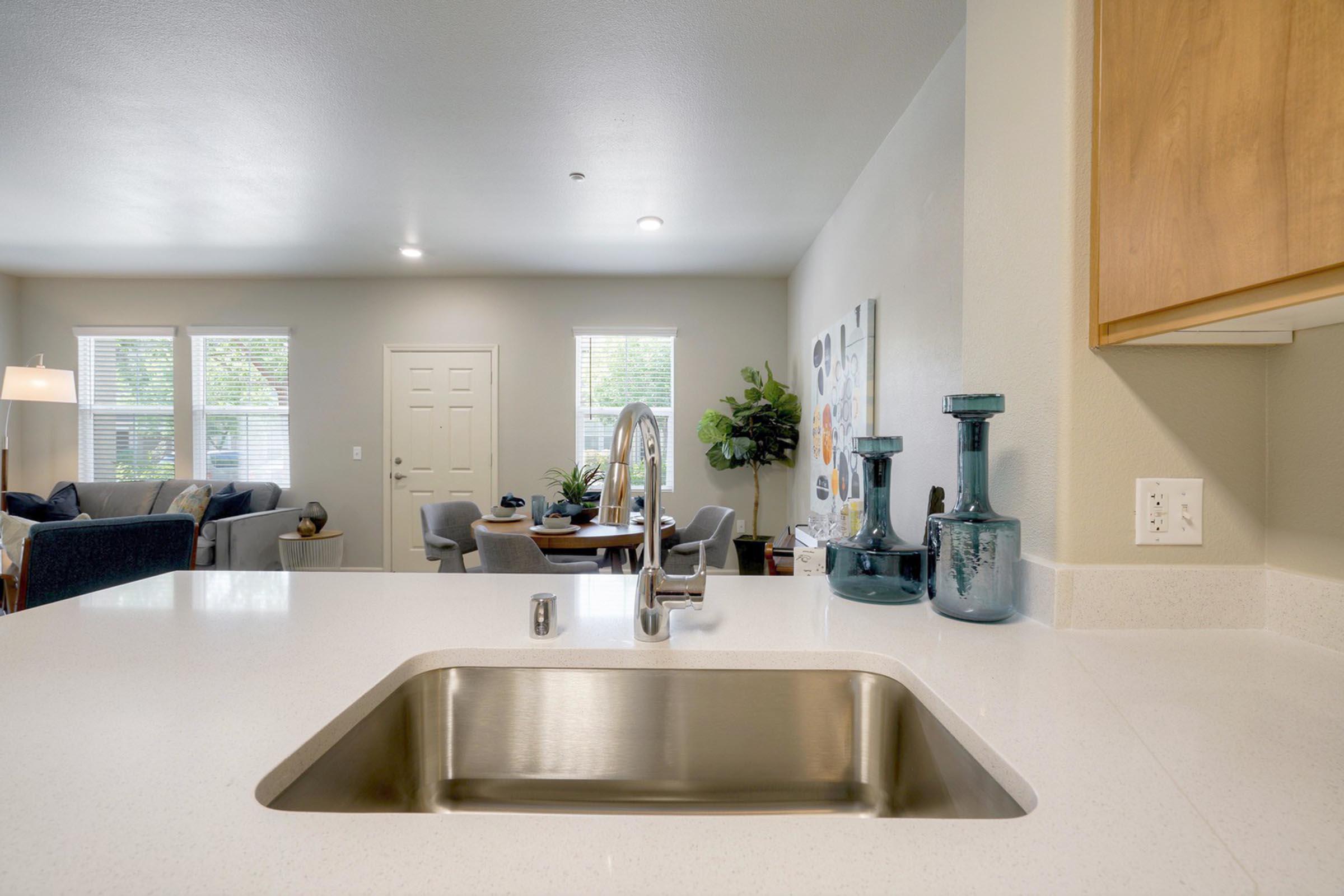 A modern kitchen with a stainless steel sink and a quartz countertop, featuring a stylish faucet. In the background, a bright living area is visible with a sofa, armchairs, and a dining table. Natural light filters through the windows, highlighting the contemporary decor and greenery.
