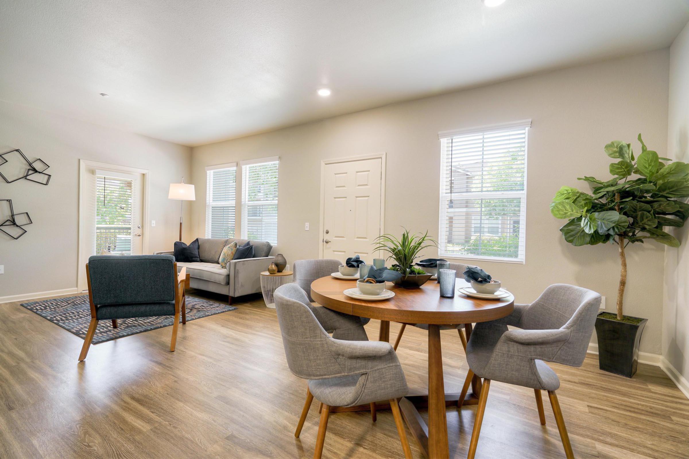 A modern living space featuring a cozy dining area with a round wooden table set for four, surrounded by grey upholstered chairs. A stylish plant stands next to the table. In the background, a comfortable sofa with cushions and a lamp is positioned beside large windows, allowing natural light to fill the room.