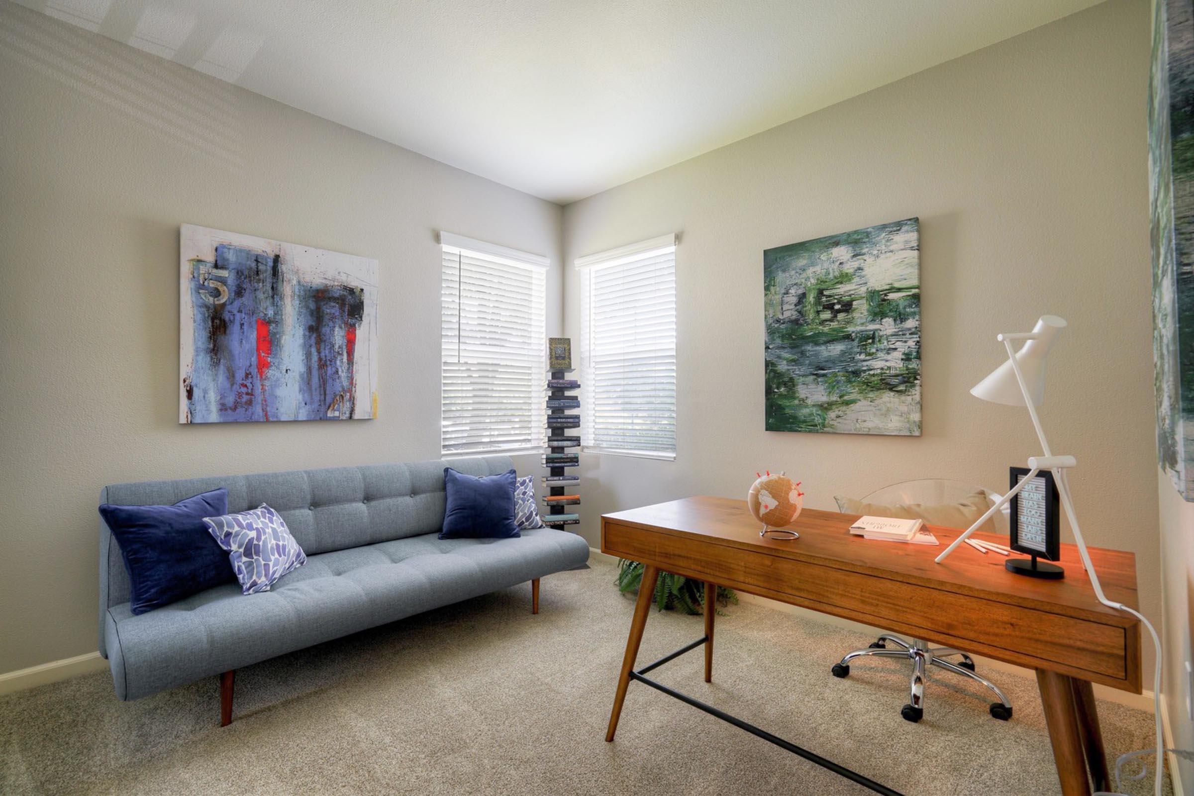A bright and modern home office featuring a light gray sofa with decorative pillows, a wooden desk with an orange clock, and a lamp. Two large windows let in natural light, and there are abstract art pieces on the walls. A tall bookcase filled with books adds to the cozy and stylish atmosphere.