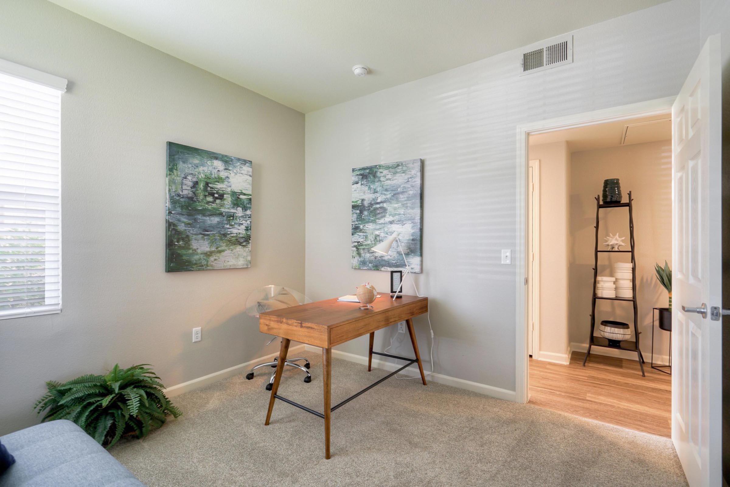 A minimalist home office featuring a wooden desk with a chair, two abstract paintings on the walls, and a small plant. The room has natural light from a window with blinds, light gray walls, and a doorway leading to another room visible in the background.