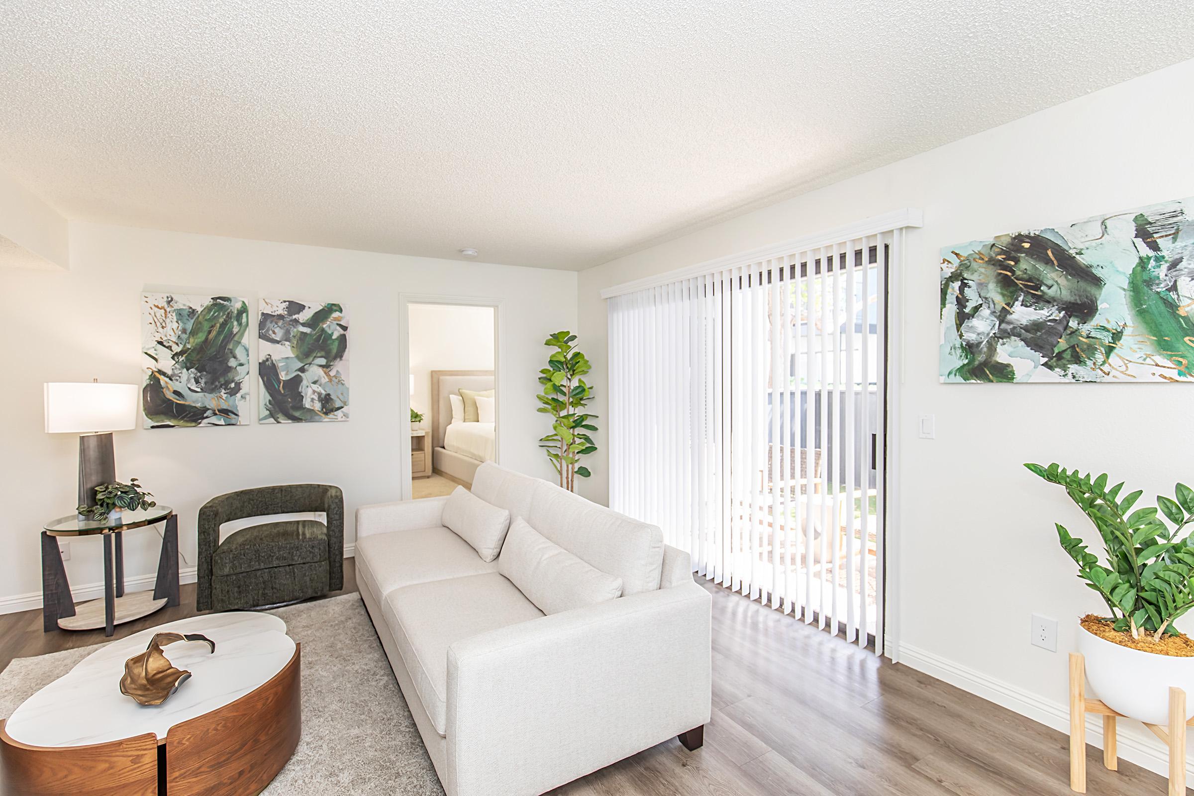 A modern living room featuring a white sofa, a round wooden coffee table, and a green accent chair. The space includes abstract artwork on the walls, a tall plant by the window, and vertical blinds allowing natural light. A doorway leads to another room in the background.