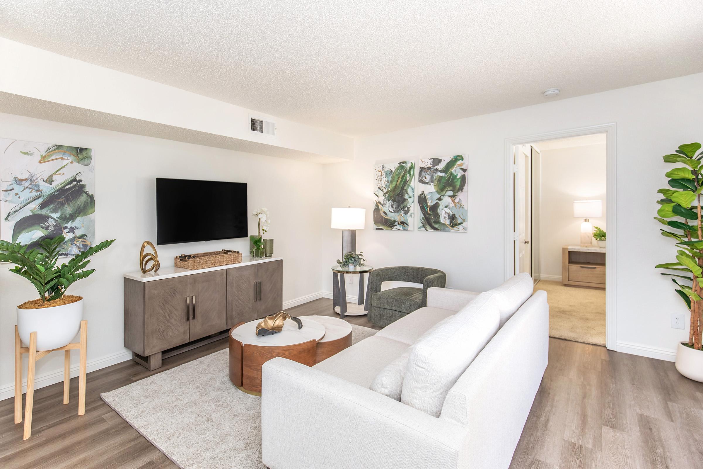 A modern living room featuring a light-colored sofa, a round coffee table with decorative items, and a TV mounted on a gray console. The walls are adorned with abstract art, and there are potted plants adding greenery. A doorway leads to another room, and soft lighting creates a welcoming atmosphere.