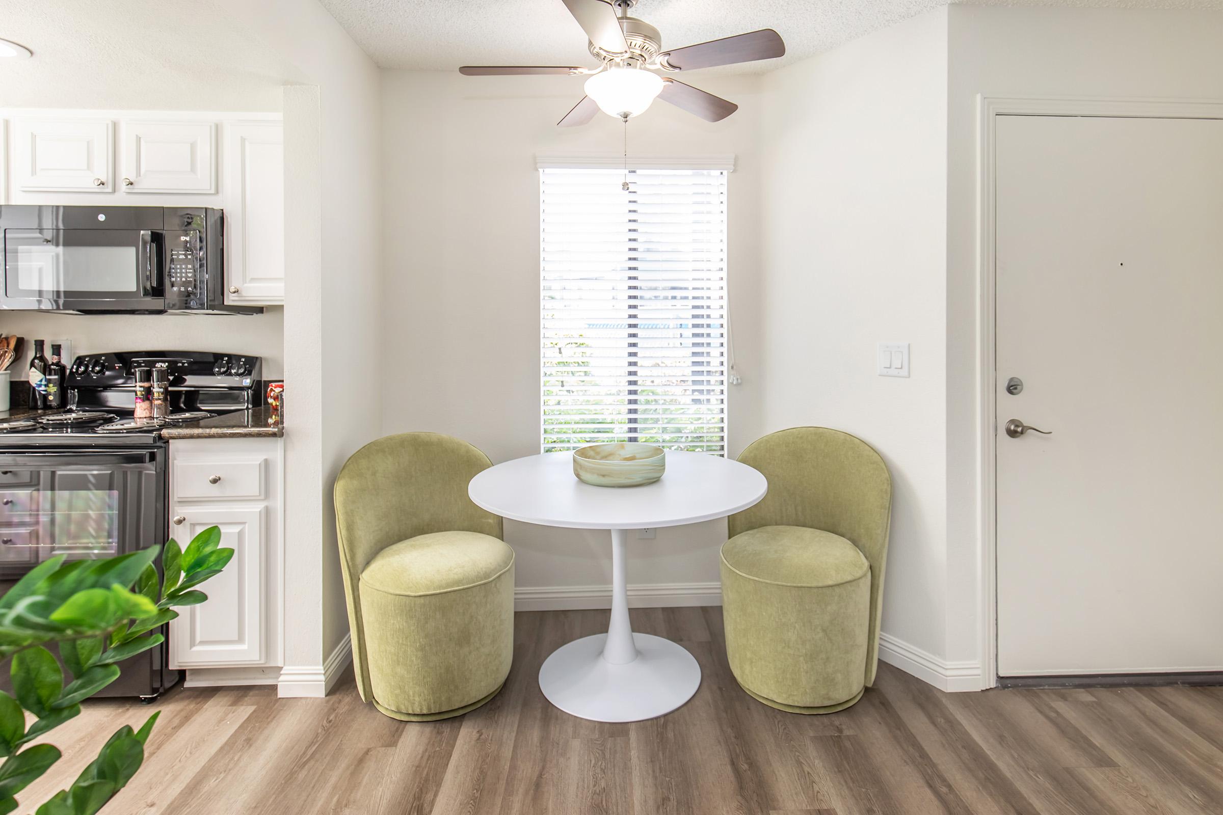 A small dining area featuring a round white table with a green vase at the center, two plush, light green chairs, and a window with white blinds letting in natural light. The kitchen space is visible in the background with white cabinets and appliances.