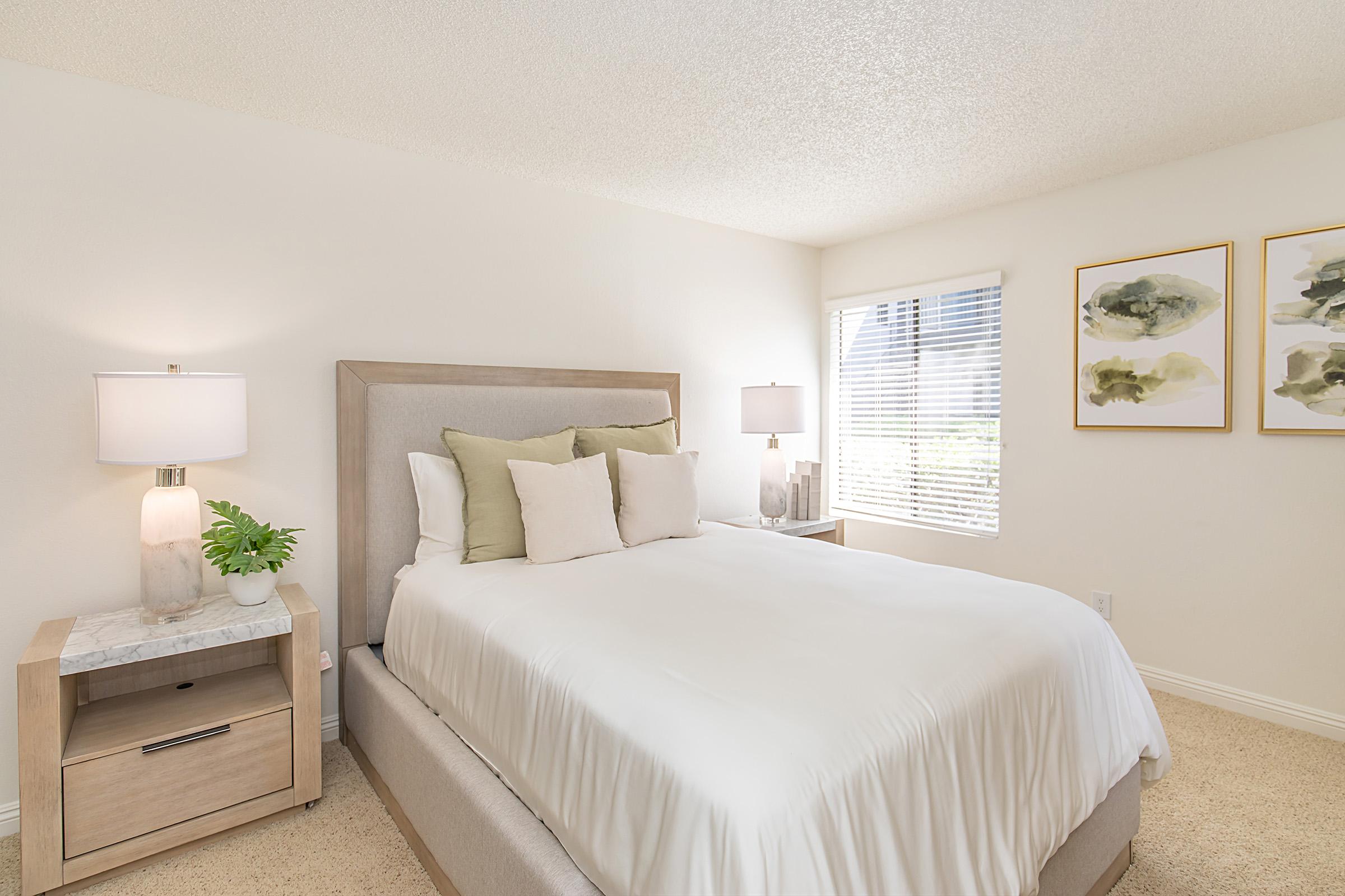 A cozy bedroom featuring a light-colored bed with white linens and decorative pillows. There are two bedside lamps on either side of the bed, along with a small nightstand. The walls are adorned with framed artwork, and a sunny window lets in natural light, creating a warm and inviting atmosphere.