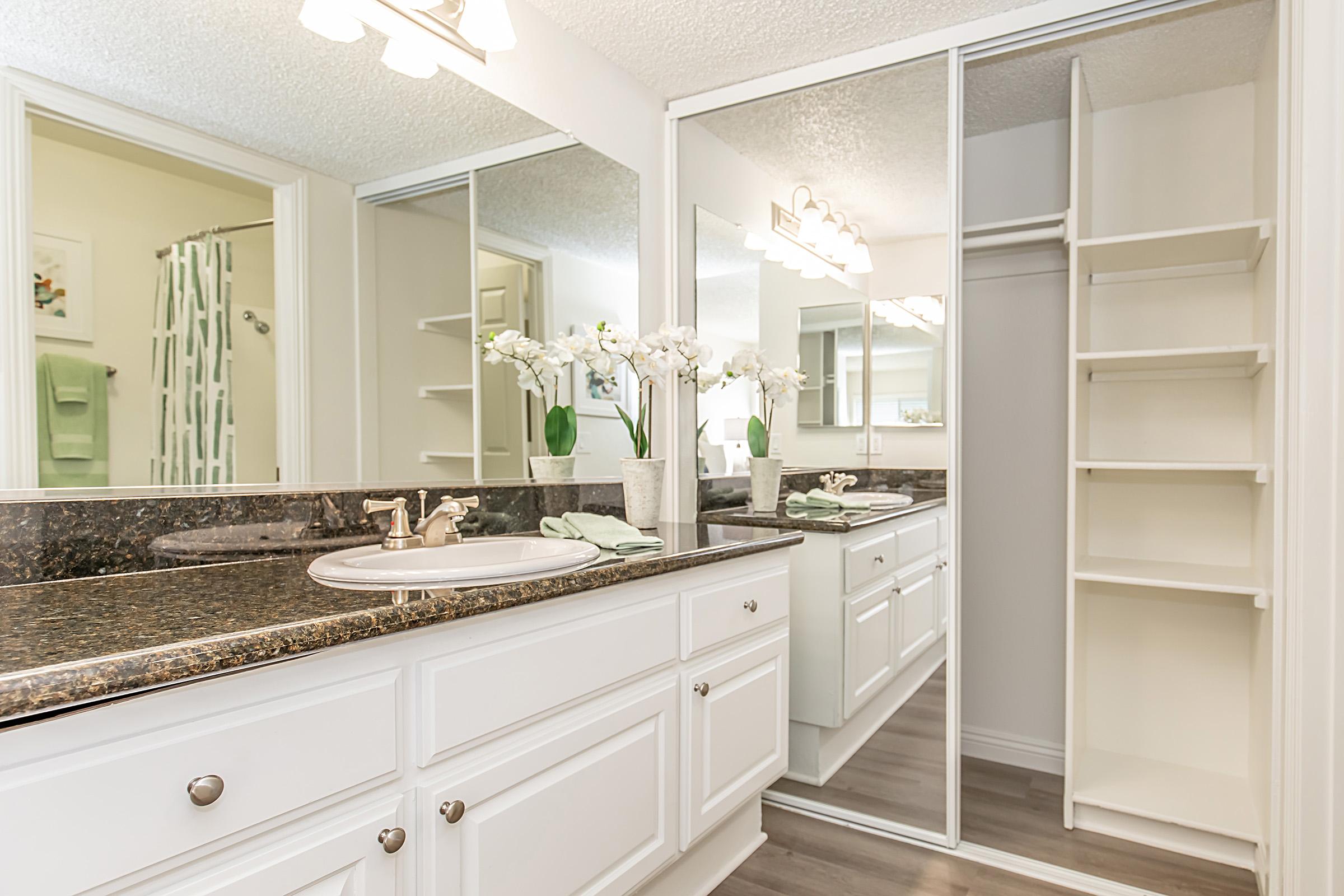 A modern bathroom featuring a double sink vanity with a dark granite countertop, framed by white cabinets. Mirrors reflect the space, creating an airy feel. Fresh white orchids in a vase add a touch of elegance, and a closet with open shelving is visible next to the vanity. Light pastel towels hang in the background.