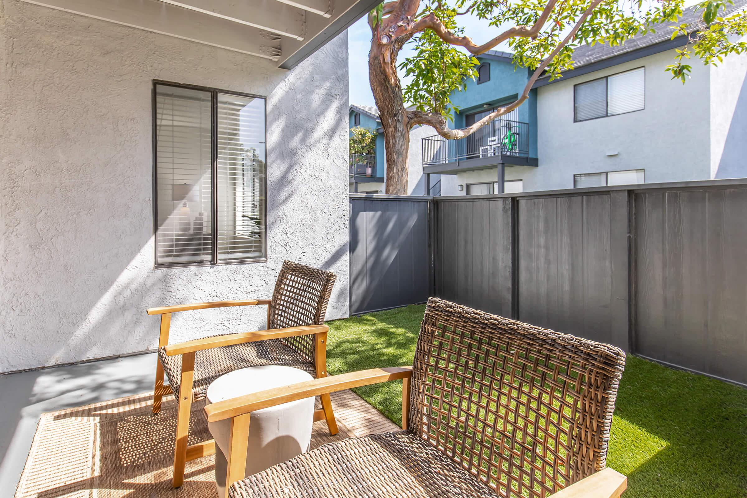 A cozy patio area featuring two wicker chairs and a small white table. Sunlight filters through the space, illuminating a well-maintained lawn and surrounding buildings with balconies. A tree adds a natural touch to the setting, creating a relaxing outdoor environment.