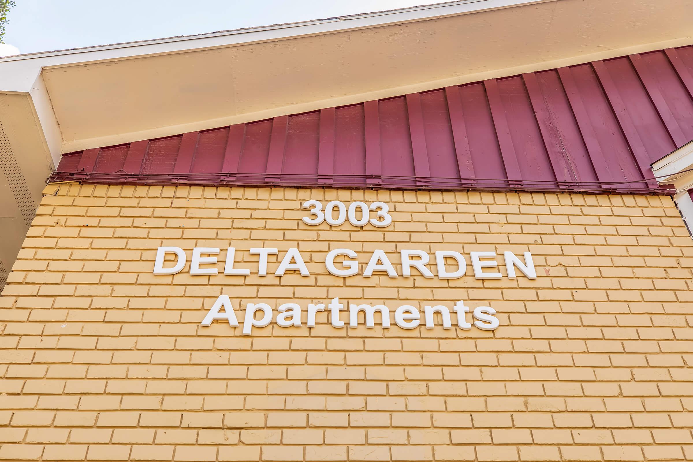 Signage for Delta Garden Apartments located at 3003, featuring a yellow brick wall and a maroon overhang. The text is prominently displayed in white letters.