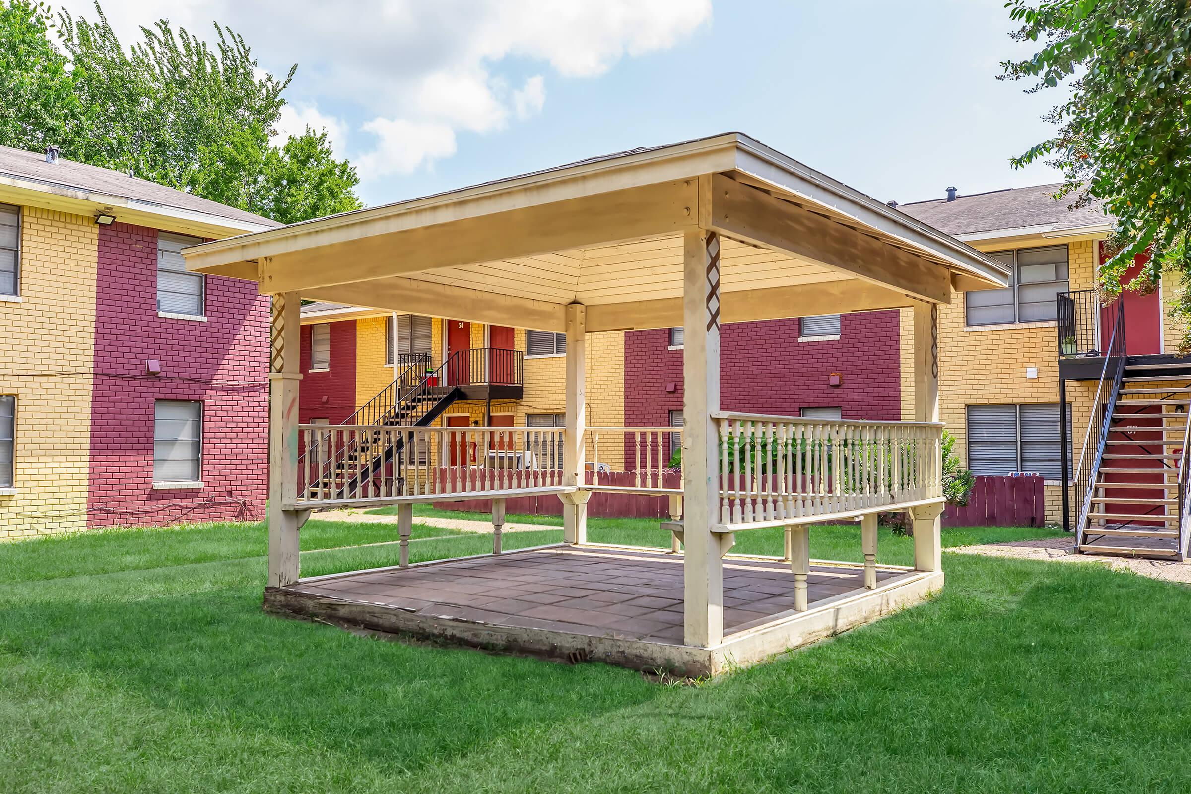 A wooden gazebo with a sloped roof and open sides stands on a grassy area, surrounded by red and yellow apartment buildings in the background. The gazebo features a flat floor and is elevated slightly above the ground, providing a shaded outdoor space. Bright blue sky and fluffy clouds are visible above.