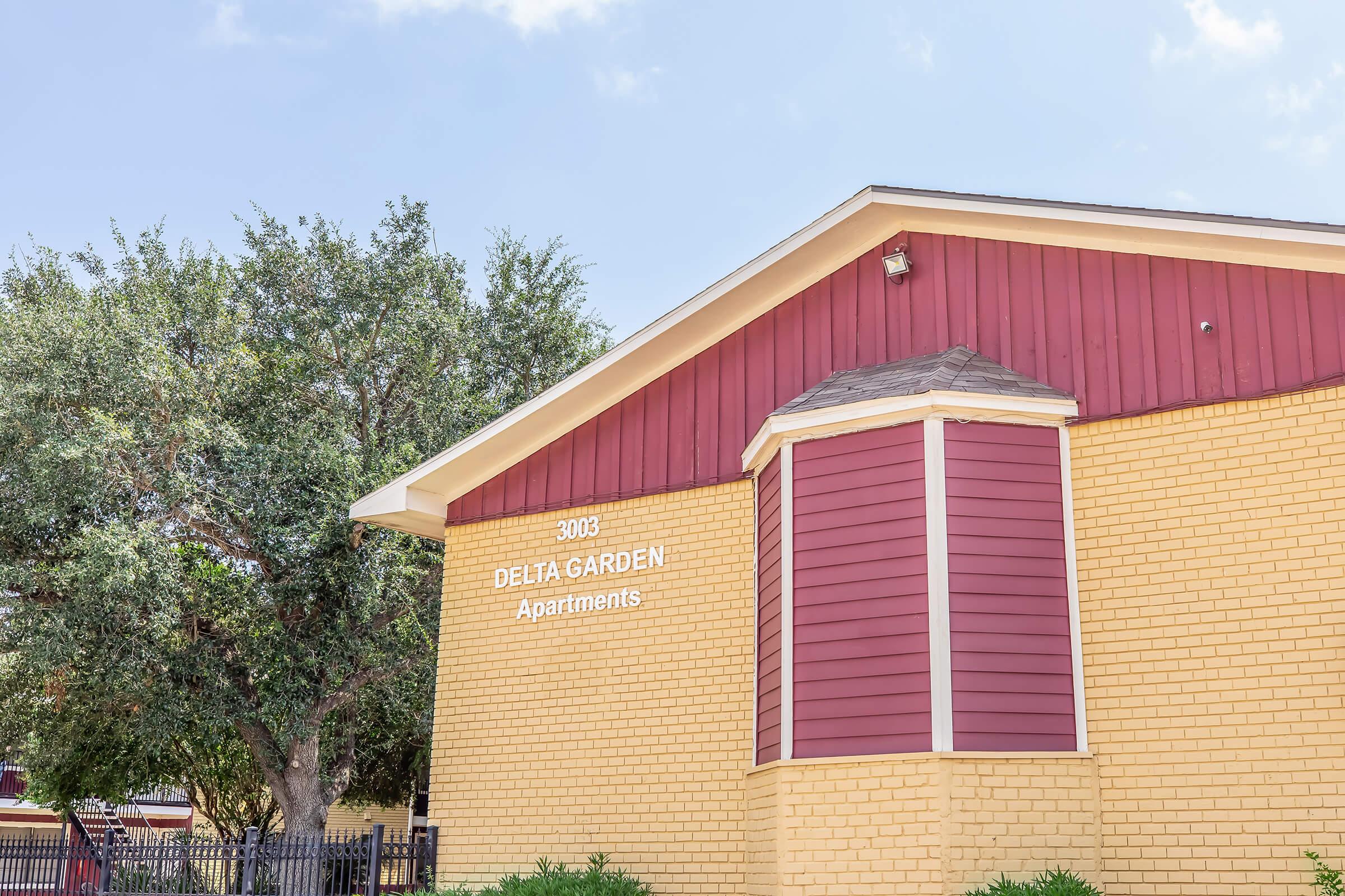 Exterior view of Delta Garden Apartments, featuring a two-tone color scheme with yellow brick and red accents. The building has a sloped roof and a sign displaying the name and address, surrounded by landscaping and trees on a sunny day.