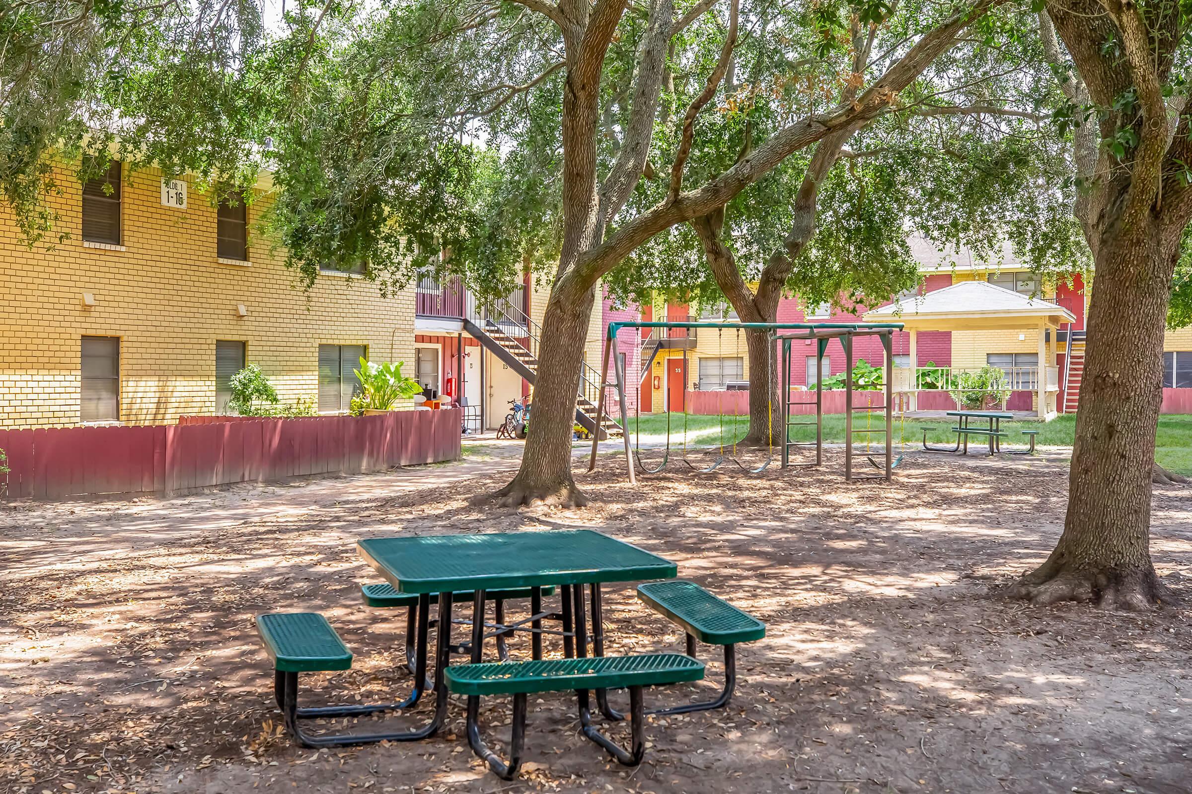 A shaded outdoor area featuring a green picnic table with attached benches, surrounded by trees and grass. In the background, there are red and yellow apartment buildings and a playground with swings. The scene is bright and inviting, perfect for relaxation or play.