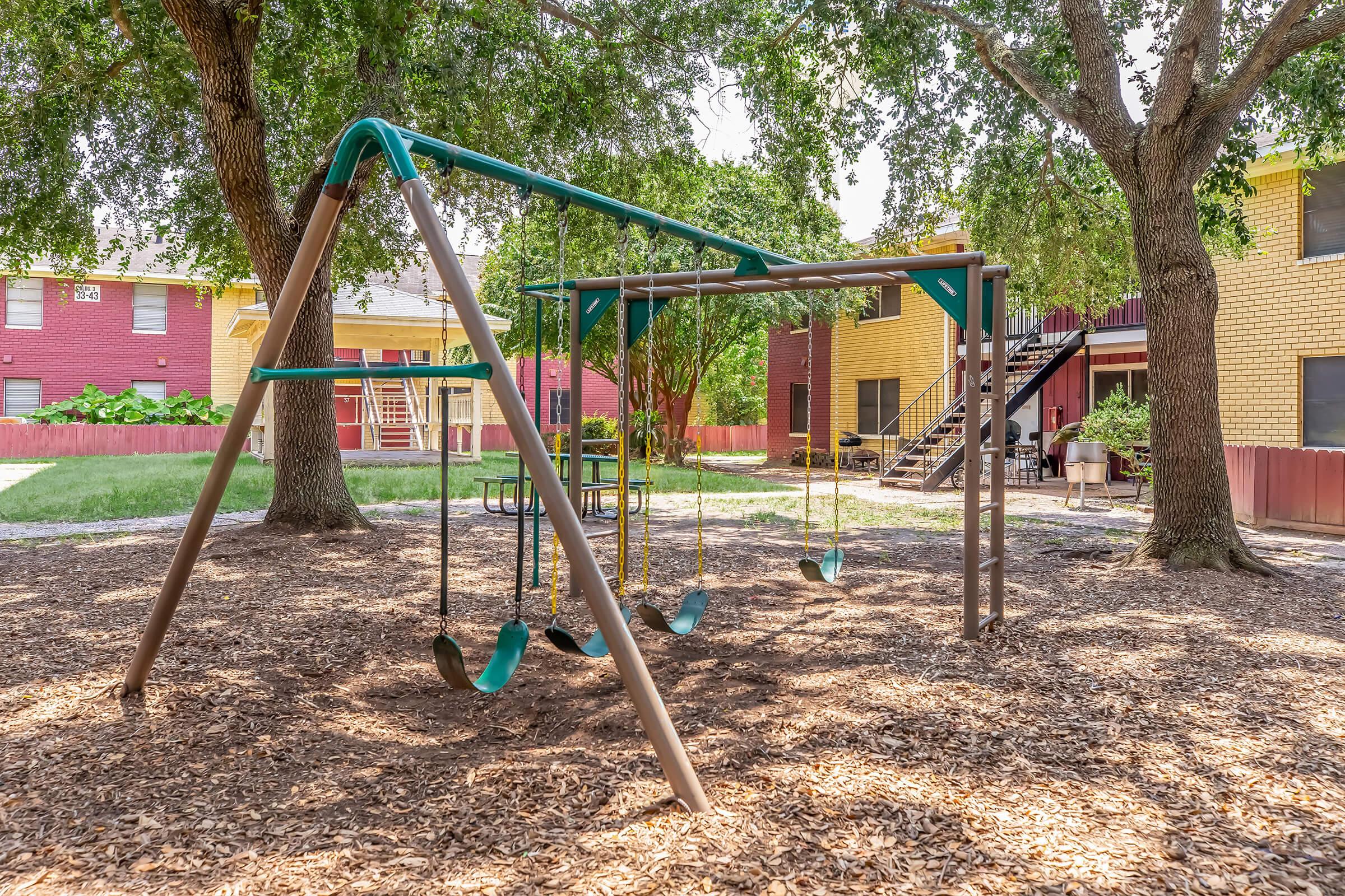 A playground featuring a swingset under two large trees, with swings hanging from a frame. In the background, there are residential buildings and a picnic table. The ground is covered with wood chips, creating a safe play area.