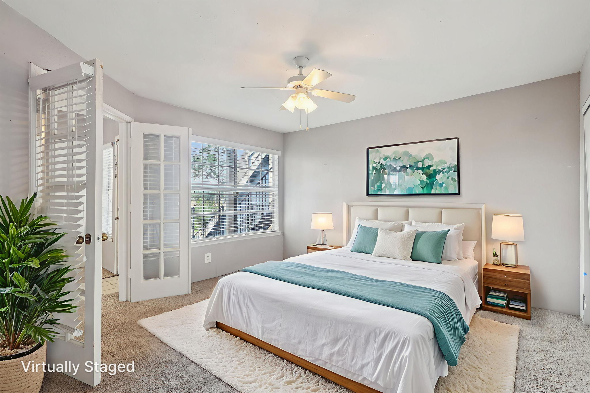 A bright and cozy bedroom featuring a large bed with white bedding and a teal throw. The room has a ceiling fan, two bedside tables with lamps, and a tropical plant. Large windows allow natural light, and a soft rug adds comfort to the space. The walls are painted a light color, creating an inviting atmosphere.