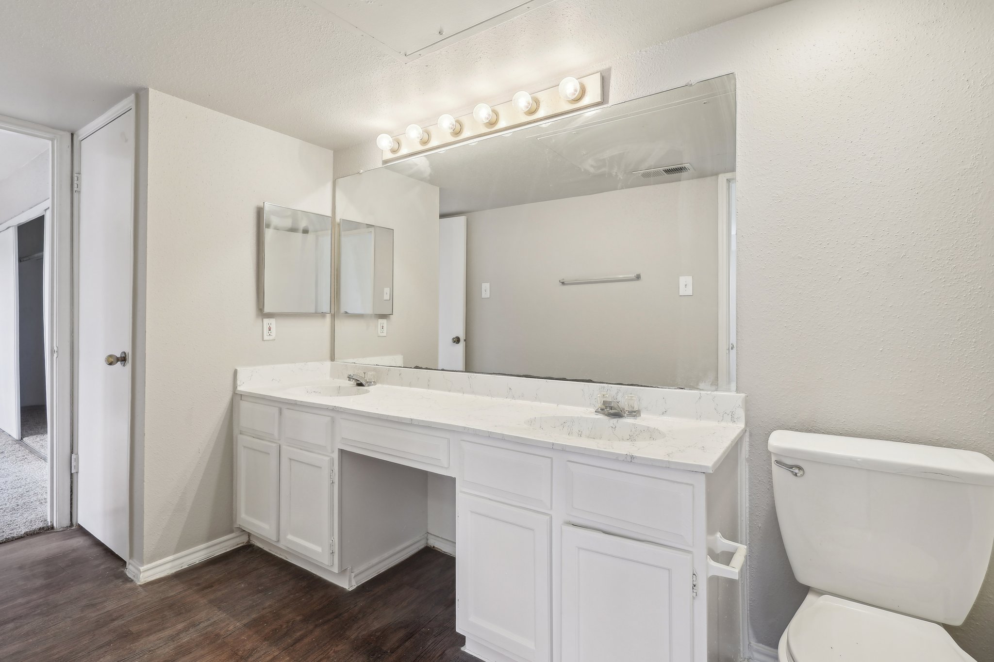 A modern bathroom featuring double sinks with marble countertops, a large mirror above, and white cabinetry. There is a toilet on the right side and a light fixture with bulbs above the mirror. The walls are painted light gray, and the floor has a wooden finish. A door leads to another room on the left.