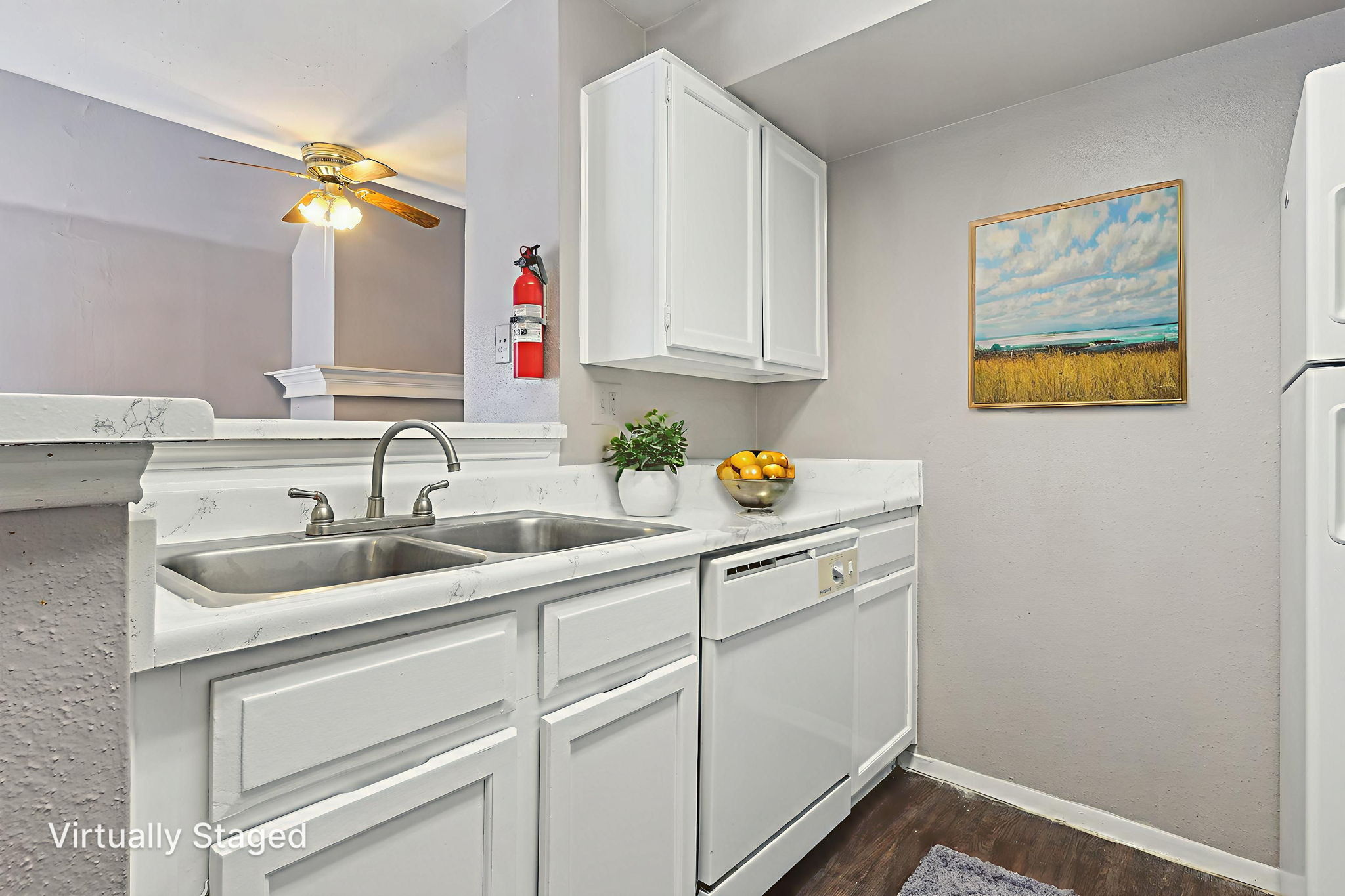 Modern kitchen with white cabinets, a stainless steel sink, and a dishwasher. A bowl of oranges is placed on the countertop, and a framed landscape painting hangs on the wall. The space features neutral walls, hardwood flooring, and a ceiling fan. Fire extinguisher mounted nearby for safety.