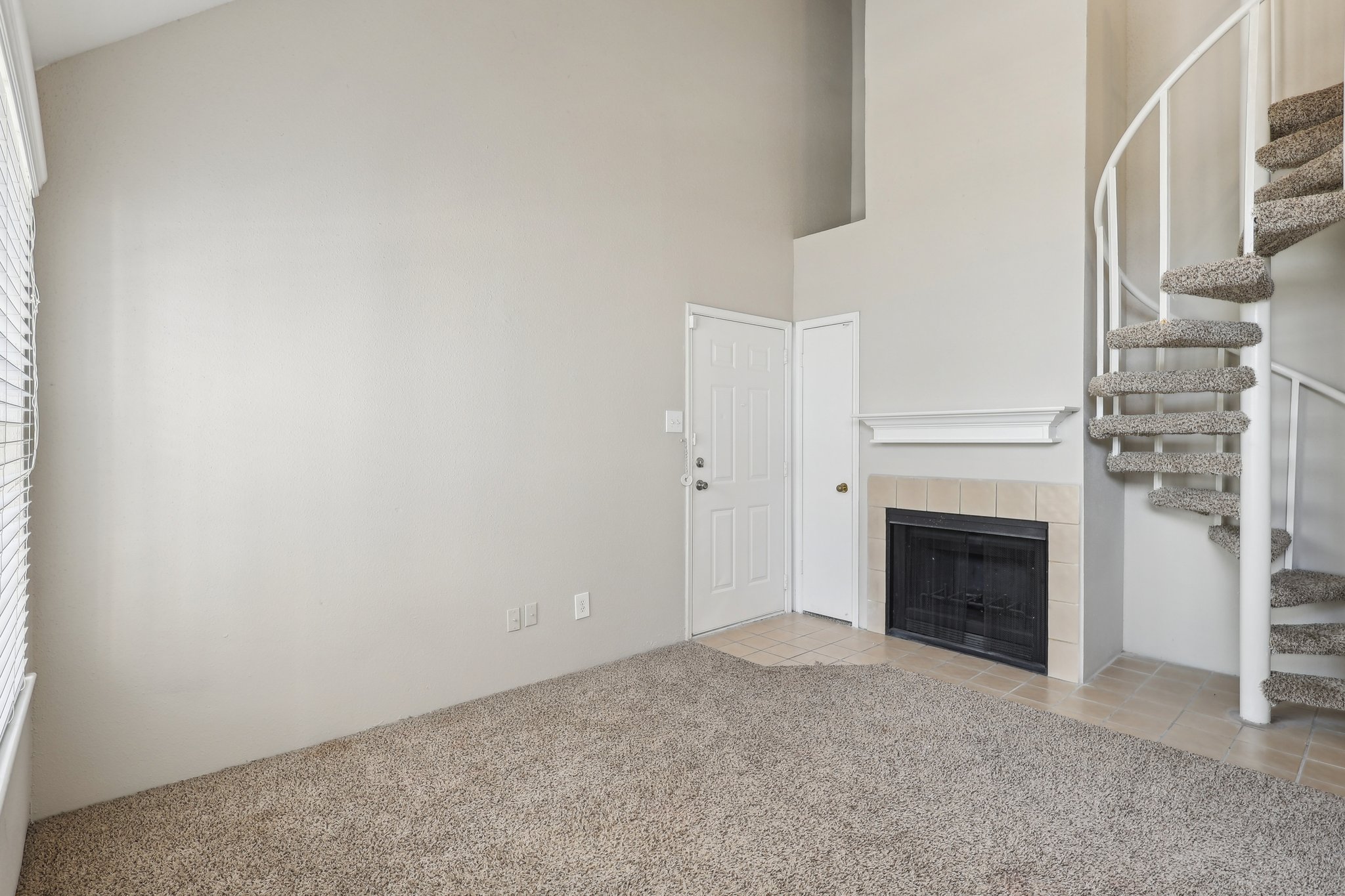 A spacious living area featuring light beige walls, a door leading outside, and a cozy corner fireplace. There is a spiral staircase on the right leading to an upper level, and the floor is covered in soft, neutral carpeting, creating a warm and inviting atmosphere. Natural light filters in through the windows.