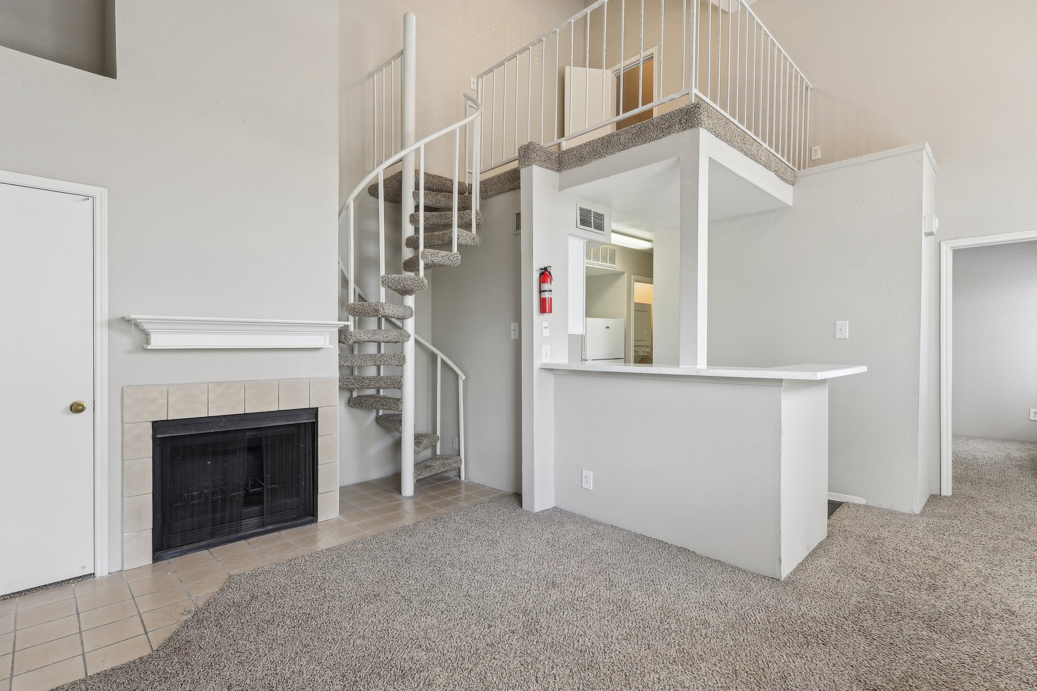 Interior view of a modern apartment, featuring a spiral staircase leading to a loft area. The room includes a fireplace, beige carpet, and a small kitchen area with a bar counter. Light-colored walls and ample natural light create a spacious feel.