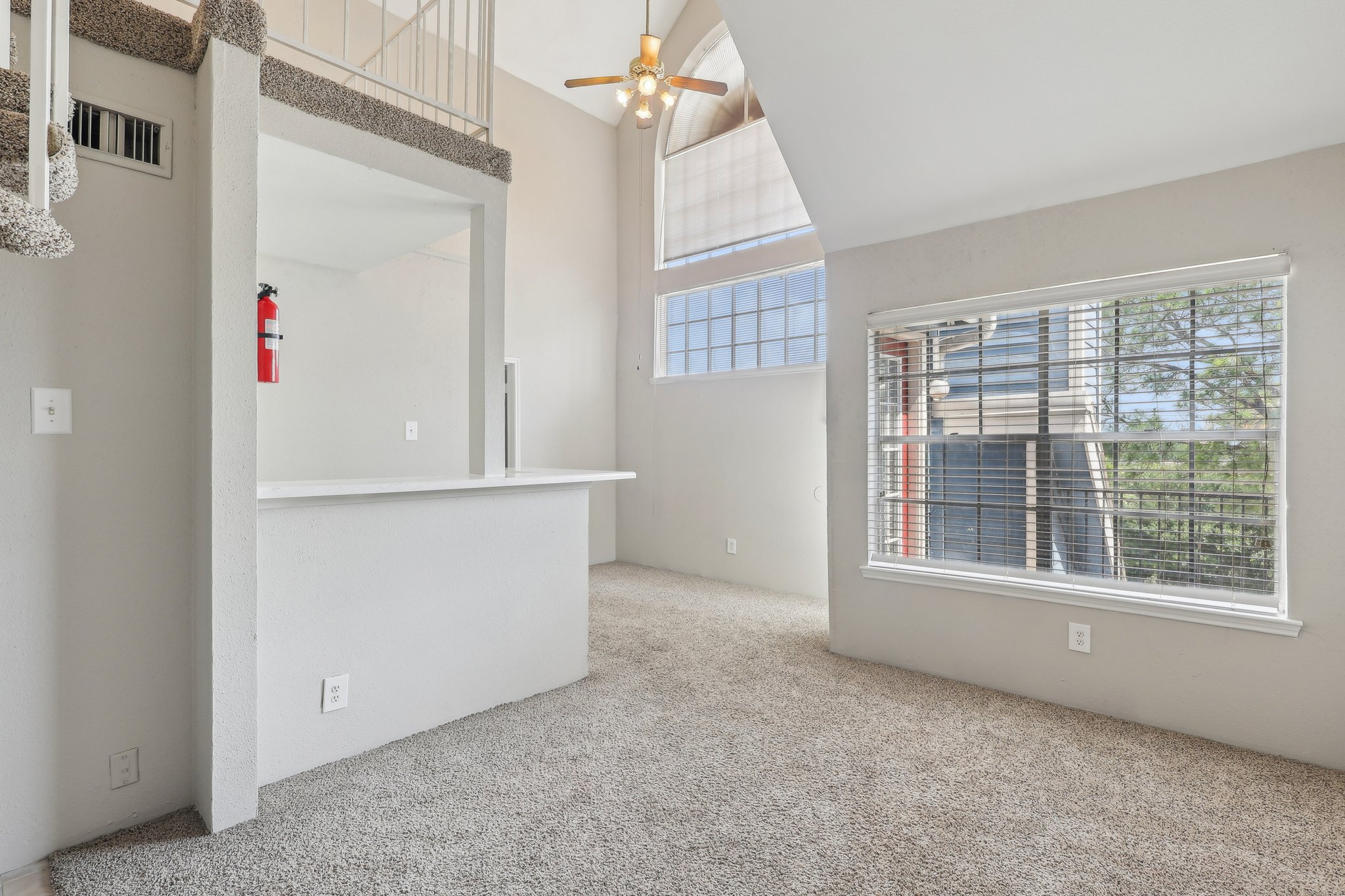 Bright and spacious room with light gray carpet, large windows allowing natural light, and a ceiling fan. A small kitchenette area is visible with a countertop. The walls are painted in neutral colors, creating an open and inviting atmosphere.