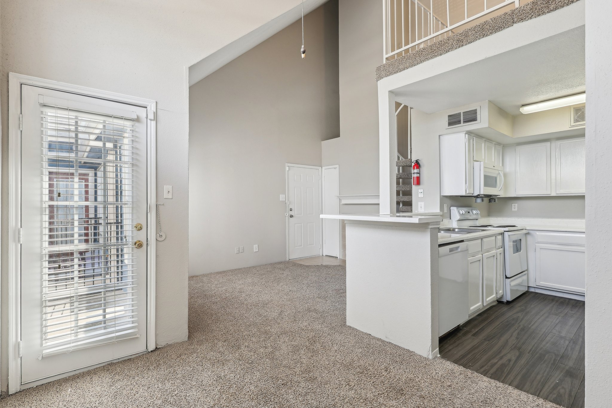A spacious interior view of an apartment featuring a living area with carpet, a kitchen with white cabinetry, and a door leading outside with blinds. The ceiling is high, and there are stairs visible leading to a second level. Bright light fills the space, creating an inviting atmosphere.