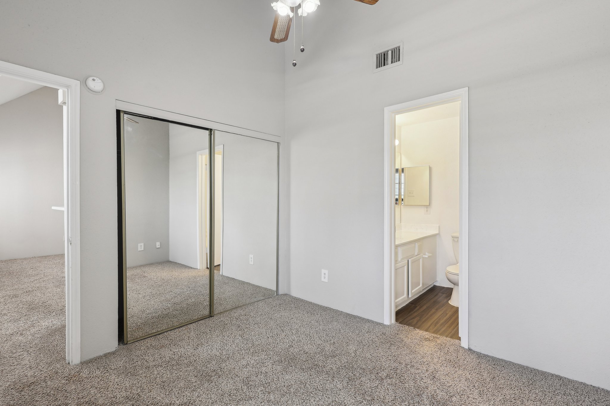 A well-lit room with gray walls and carpeted flooring. There is a mirrored closet on one side and an open doorway leading to a bathroom on the opposite side. The bathroom features a sink and mirror. Ceiling fan visible, adding to the airy feel of the space.