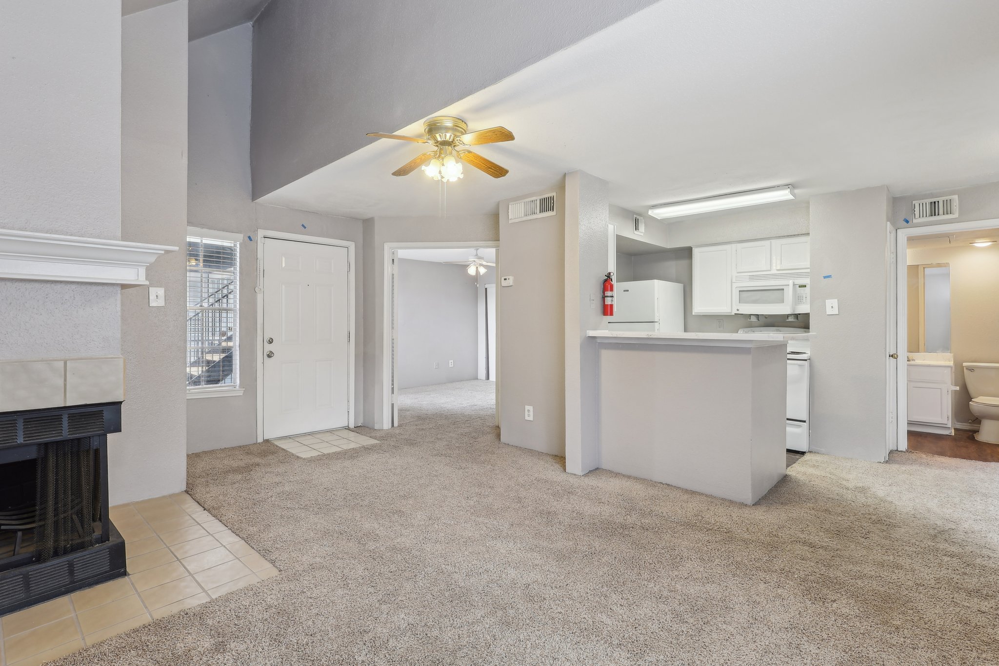 Interior view of a living space featuring a carpeted floor, a ceiling fan, a fireplace, and access to a kitchen area with white appliances. A door leads outside, and there's a doorway to a bathroom in the background. The walls are painted in soft gray tones, creating a warm and inviting atmosphere.